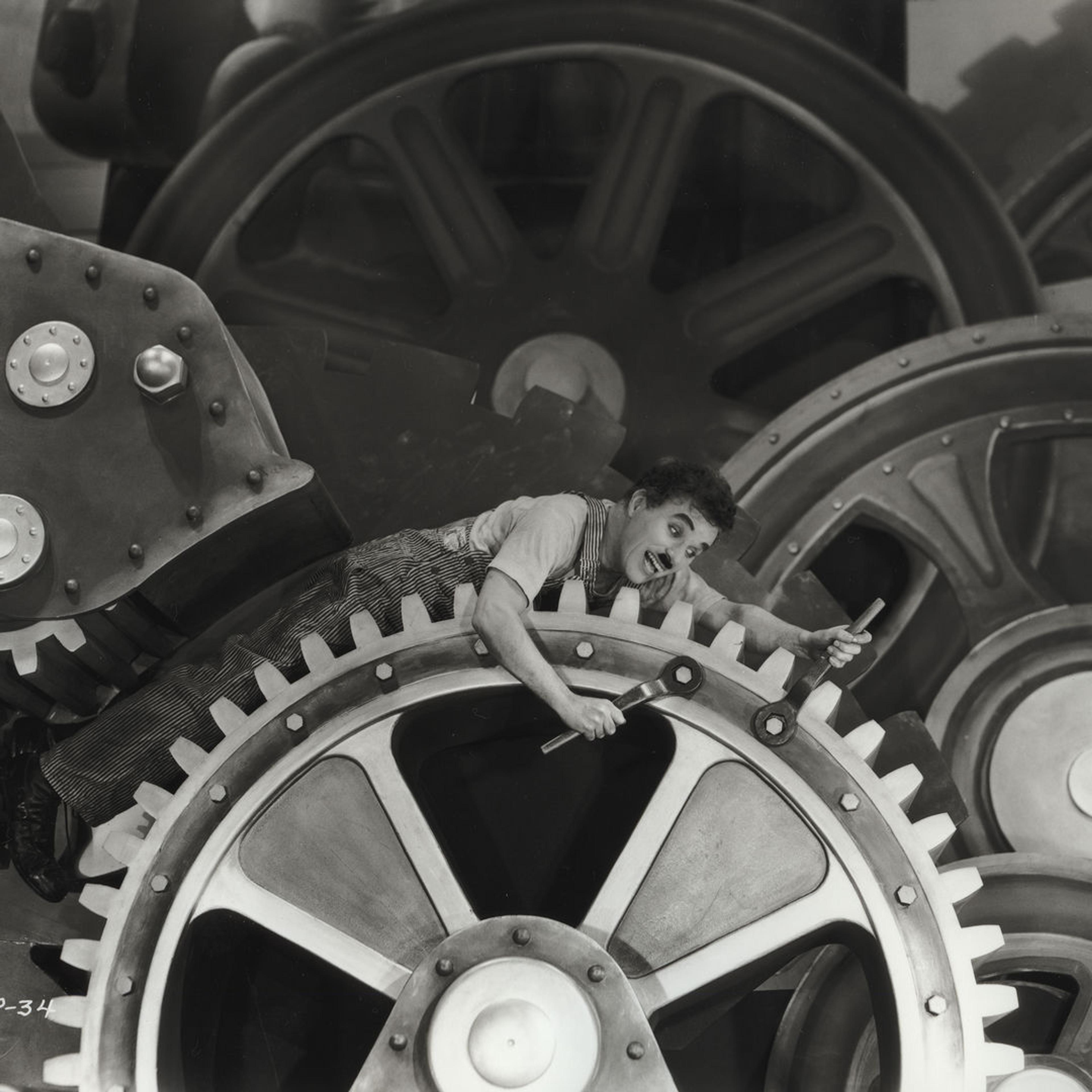 Black and white image of the actor Charlie Chaplin dressed as the tramp, a white man in overalls in this scenario and a mustache where he is trapped between massive factory cogs that he is riding while also trying to tighten the bolts with both of his hands at the same time.