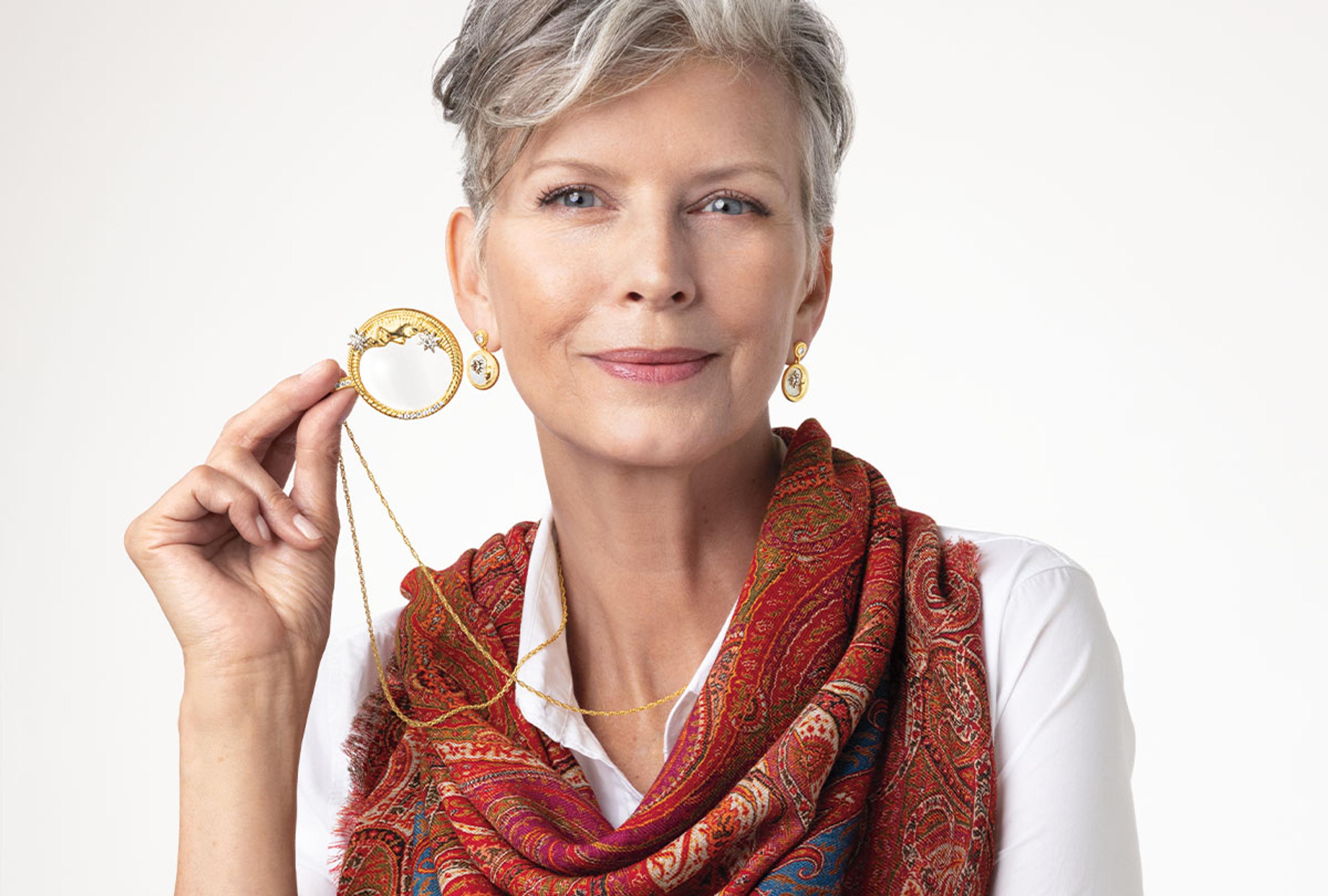 A light-skinned white woman with gray hair smiles and poses with various Met Store products including a scarf, earrings, and a necklace. 