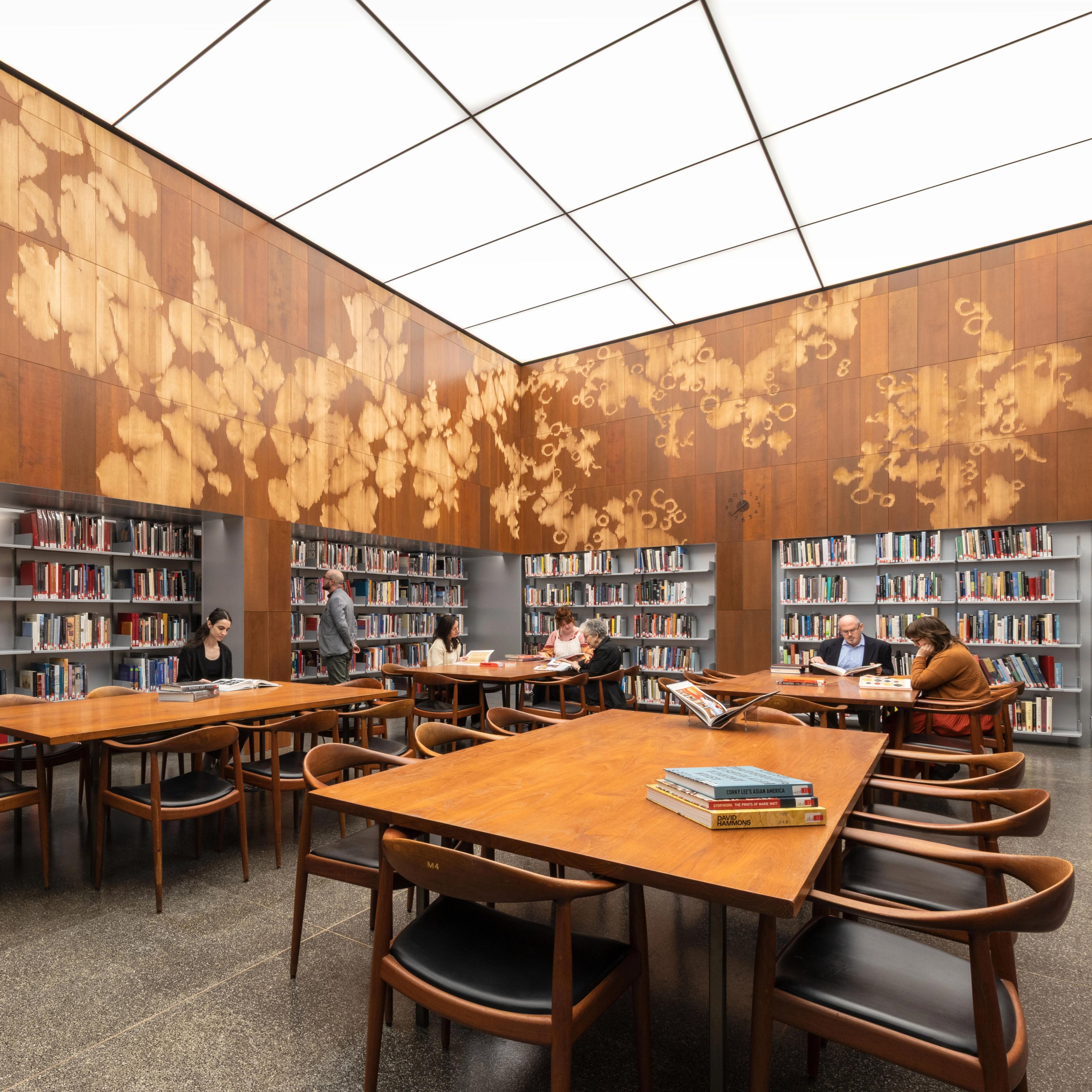 Brightly lit reading room with long tables, walls of book shelving, and wood-paneled walls with abstract motifs.