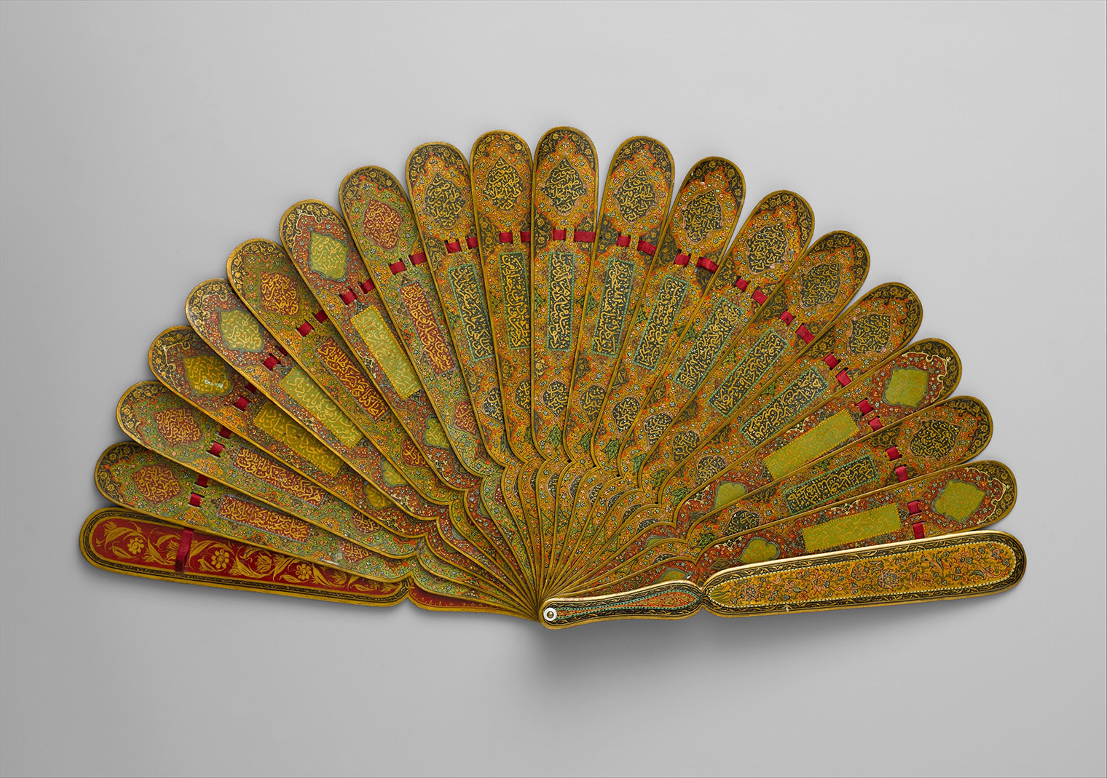Ornate gold fan with intricate designs and floral patterns, accented in red and green hues, spread out against a neutral gray background.