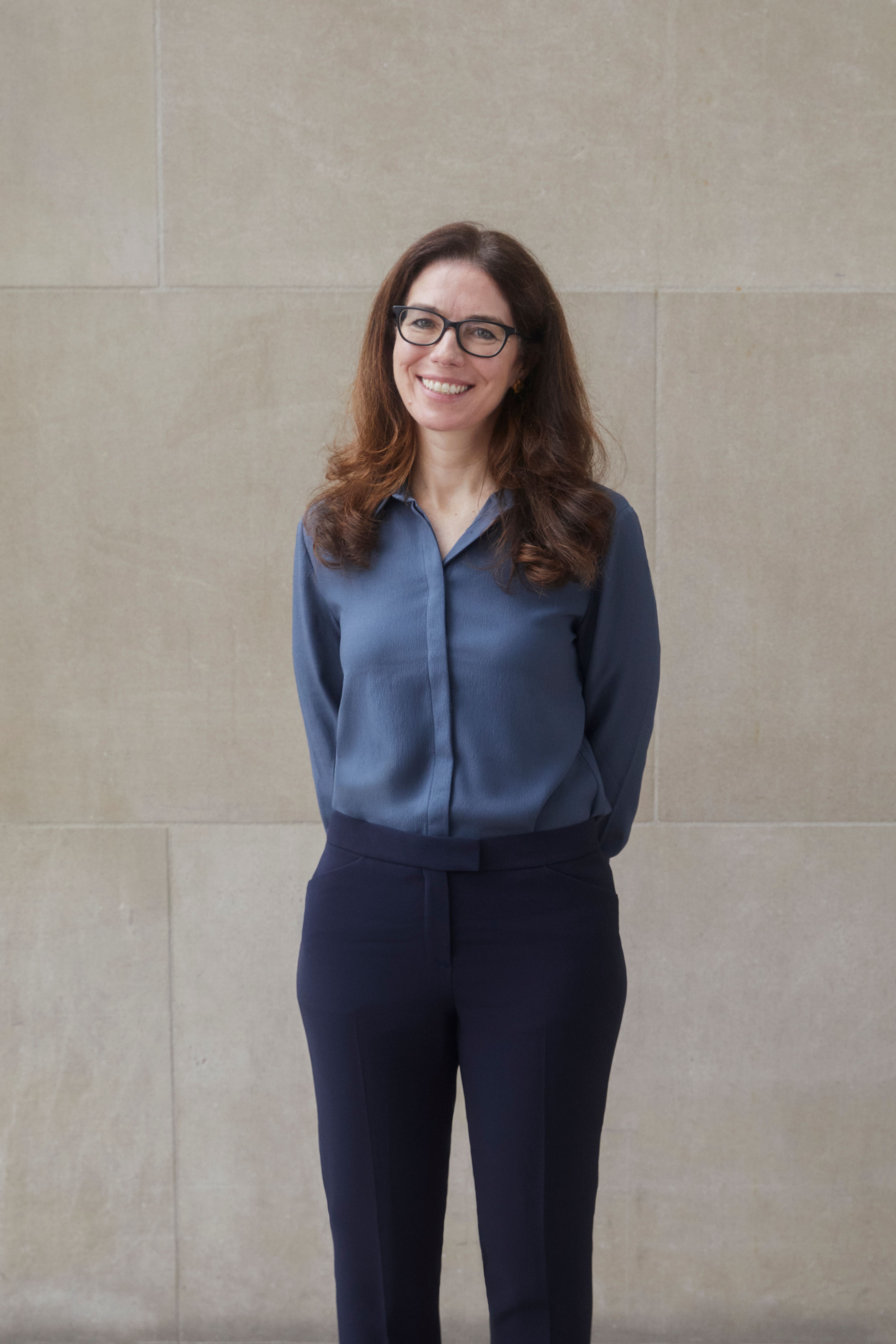 A color photograph of Jamie Kelleher standing in front of a stone wall in a blue top and black pants.