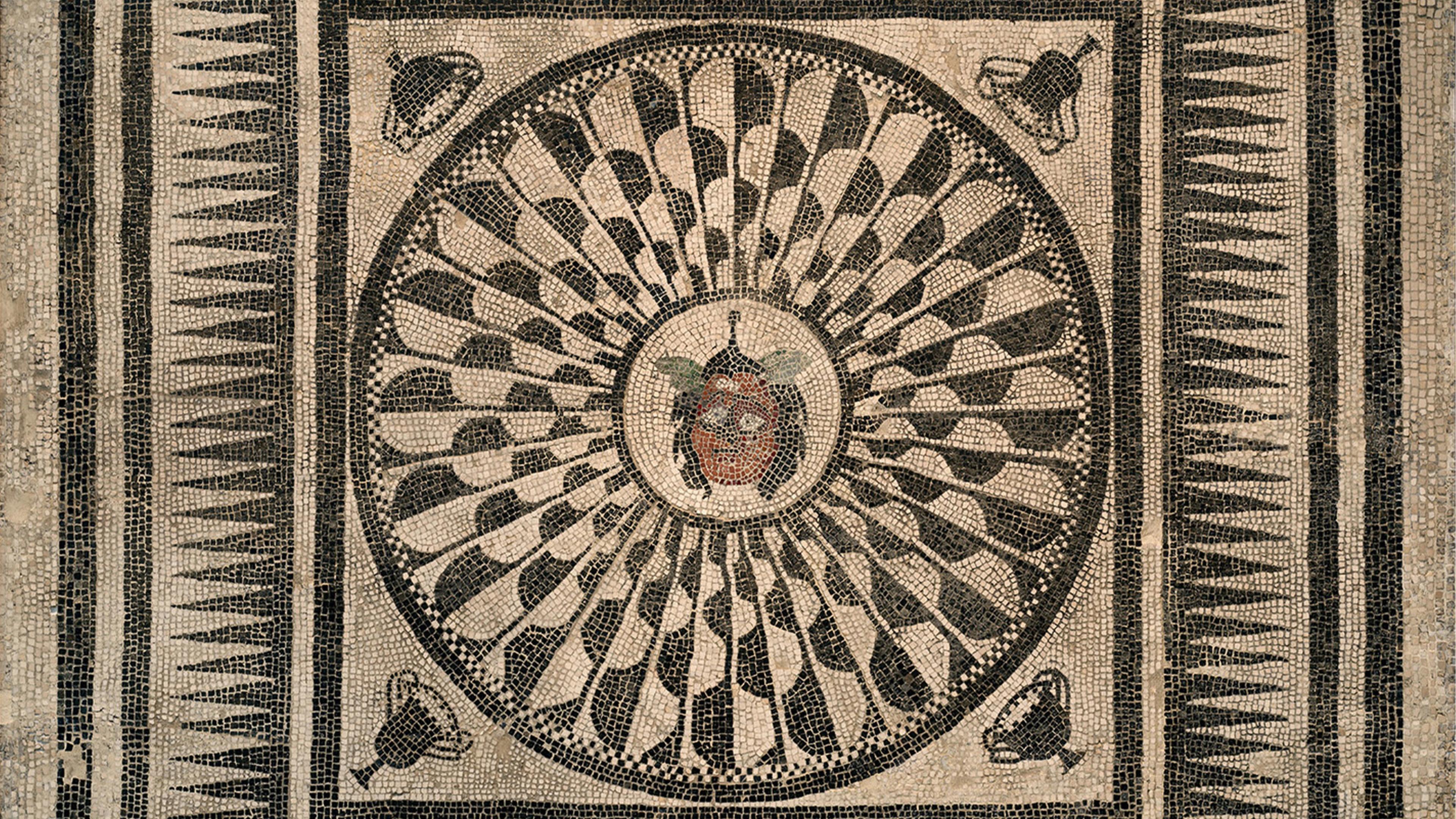 Ancient mosaic featuring a central Medusa head, radiating petal-like patterns, geometric borders, and four corner masks on a stone background.