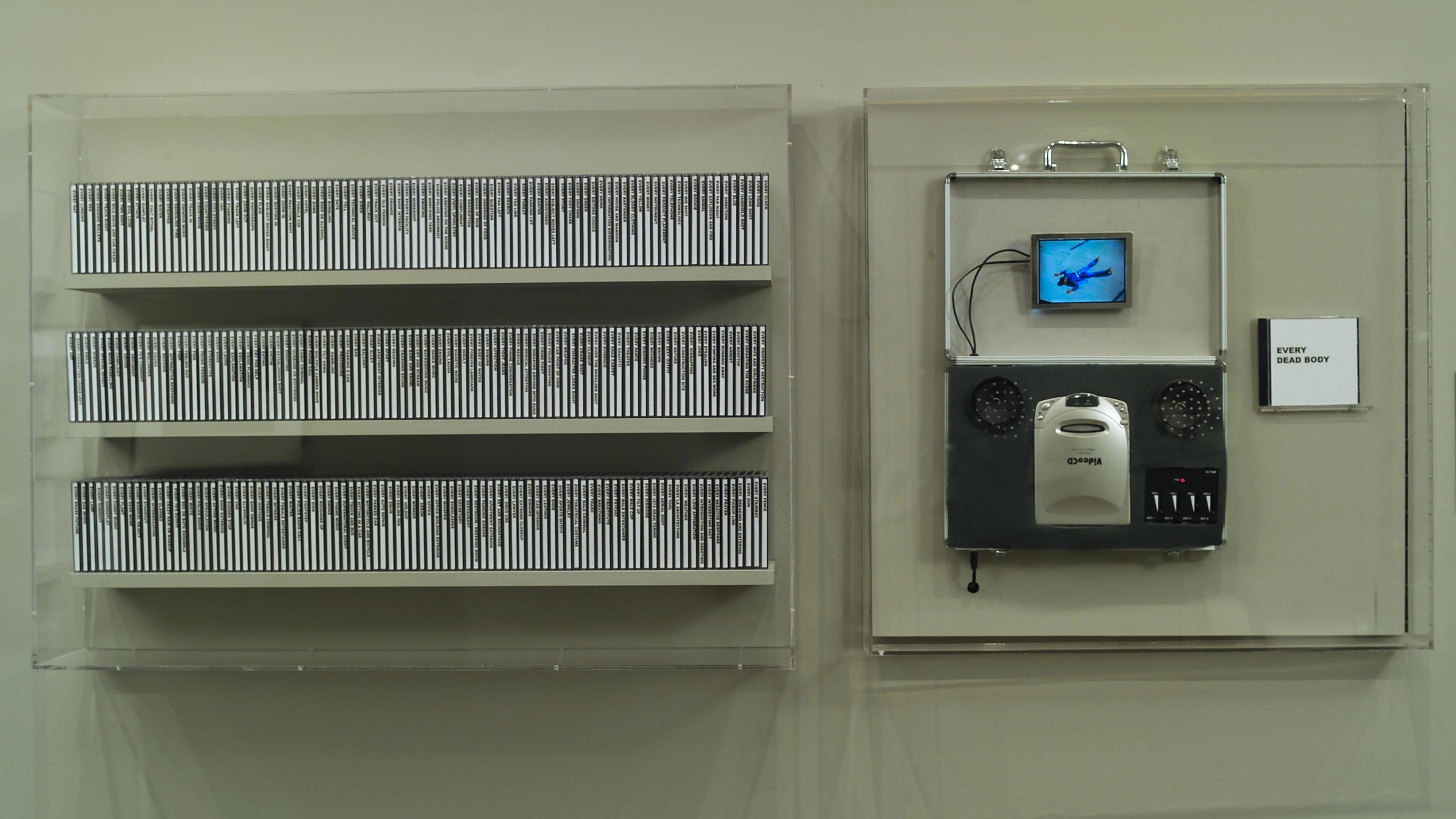 Custom digital video playback installation with 277 video compact discs displayed on three shelves next to a plexiglass case with an open suitcase containing a small screen and a VCD player. A VCD labeled "Every Dead Body" is displayed to the right of the player.