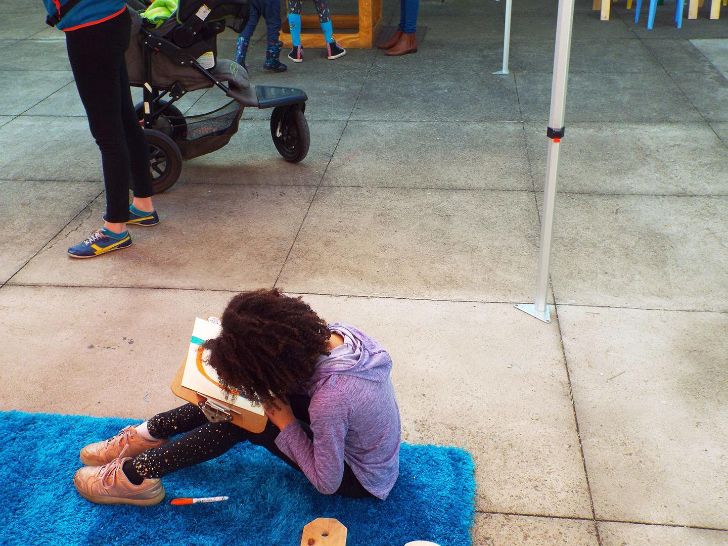 Awra is seated on the floor a shaggy blue rug. She bends over her drawing, her afro covering up most of the clipboard.