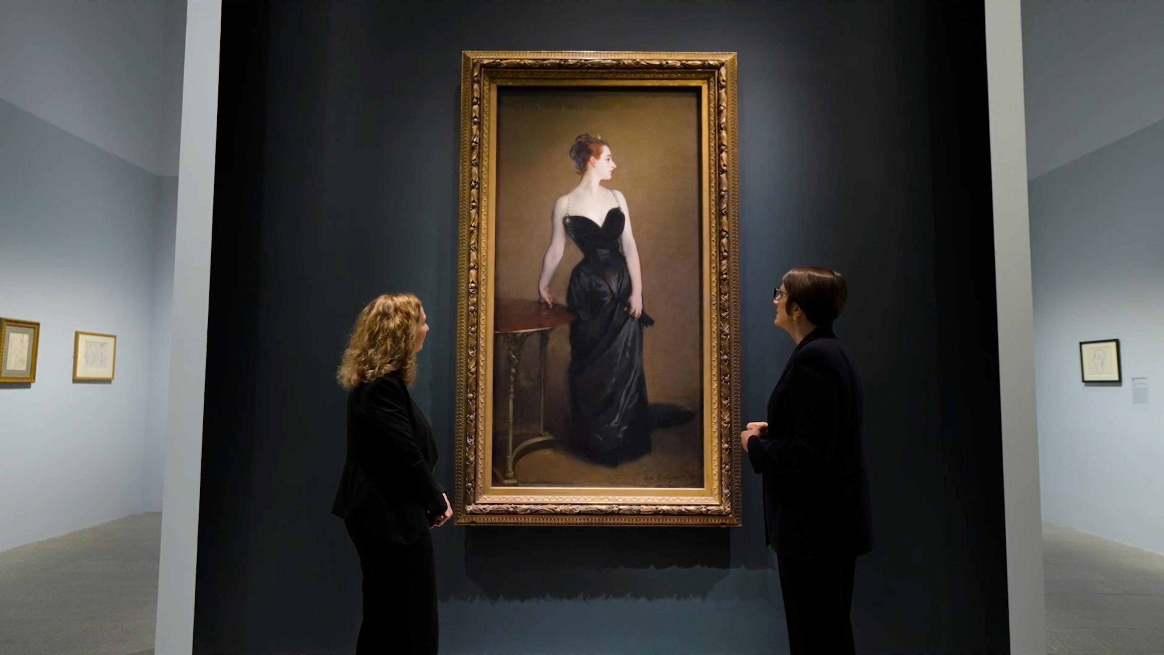 Two people admire a framed portrait of a woman in a black gown against a dark backdrop. The gallery setting is elegant and contemplative.