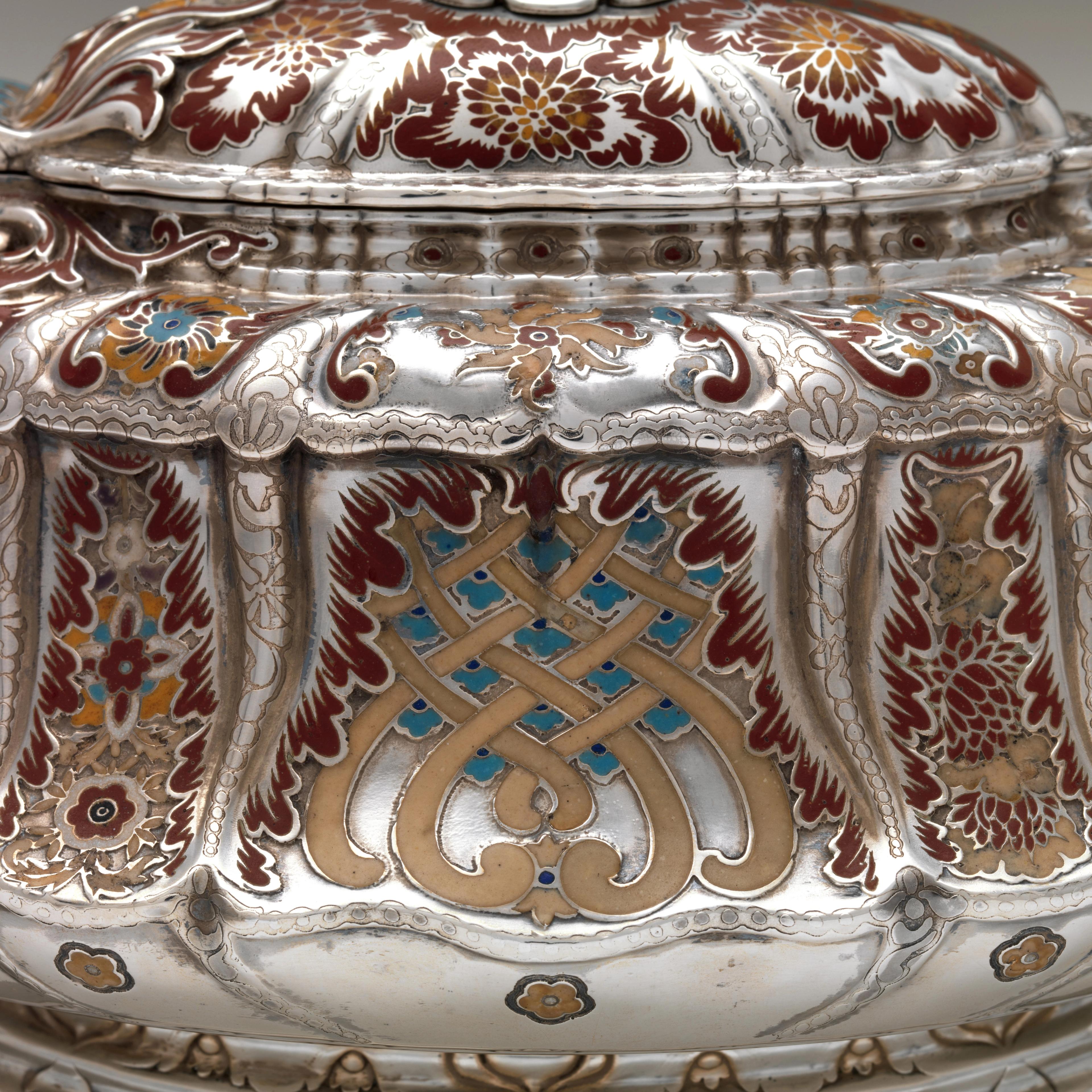 A short but elongated silver teapot with an ivory lid finial adorned with abstract floral motifs rendered in maroon, orange, taupe, and turquoise enamel.