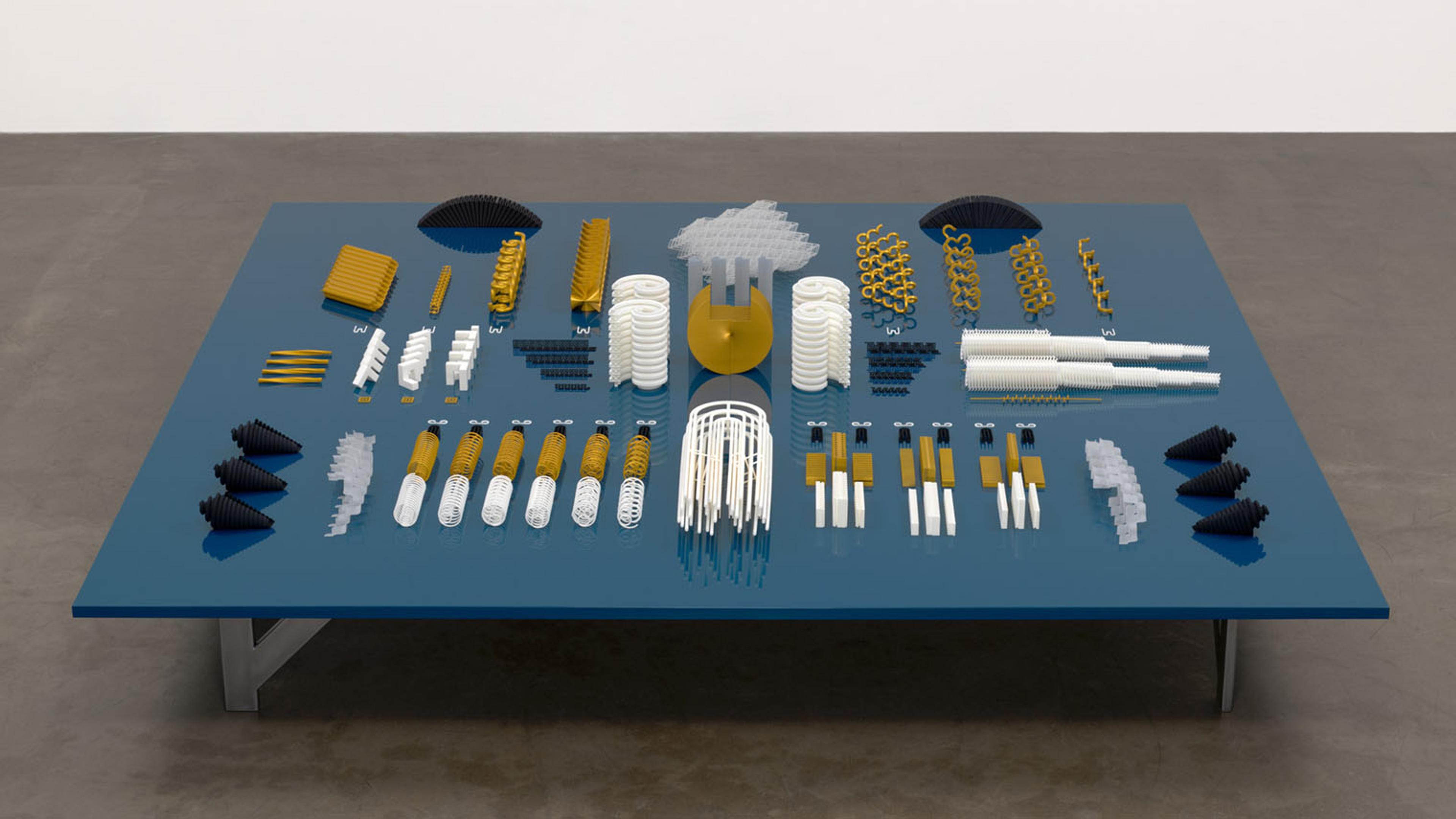Symmetrical arrangement of geometric sculptural pieces in gold, white, and black on a blue table, viewed from above.