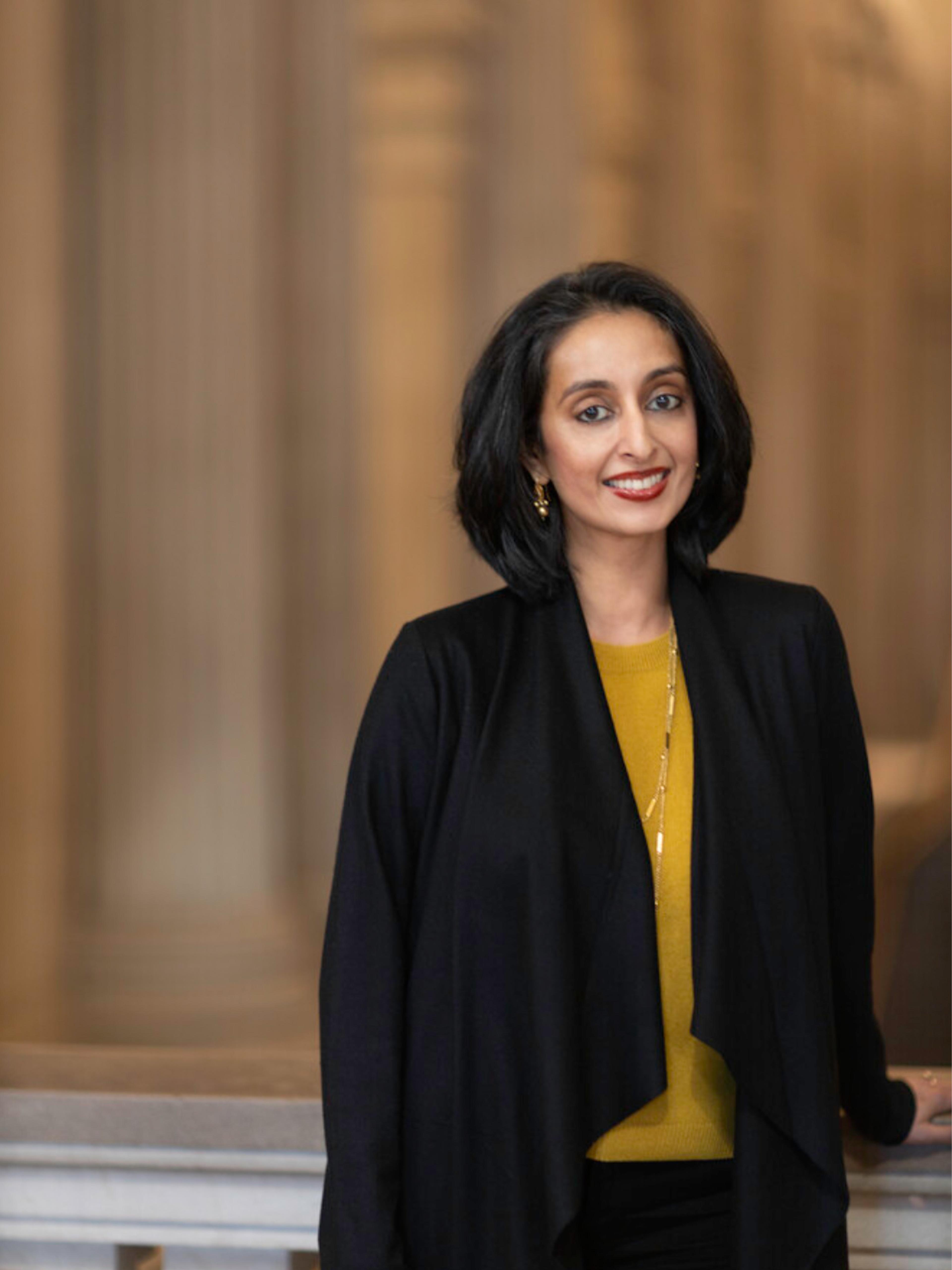 A smiling woman with short dark hair stands in a grand interior, wearing a black jacket over a mustard-yellow top.