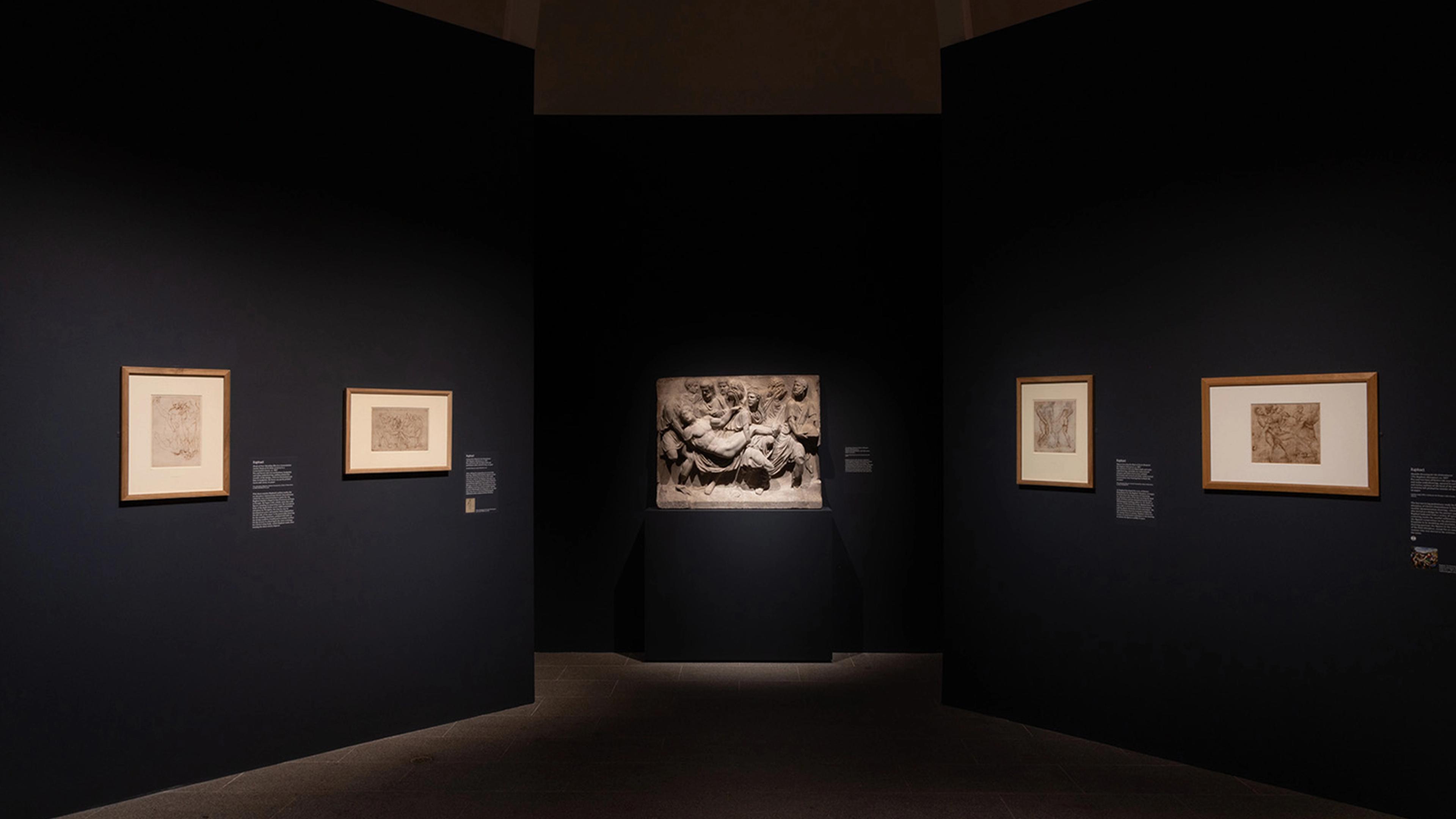 Dimly lit art gallery with dark walls features three framed sketches and a centered illuminated stone relief.