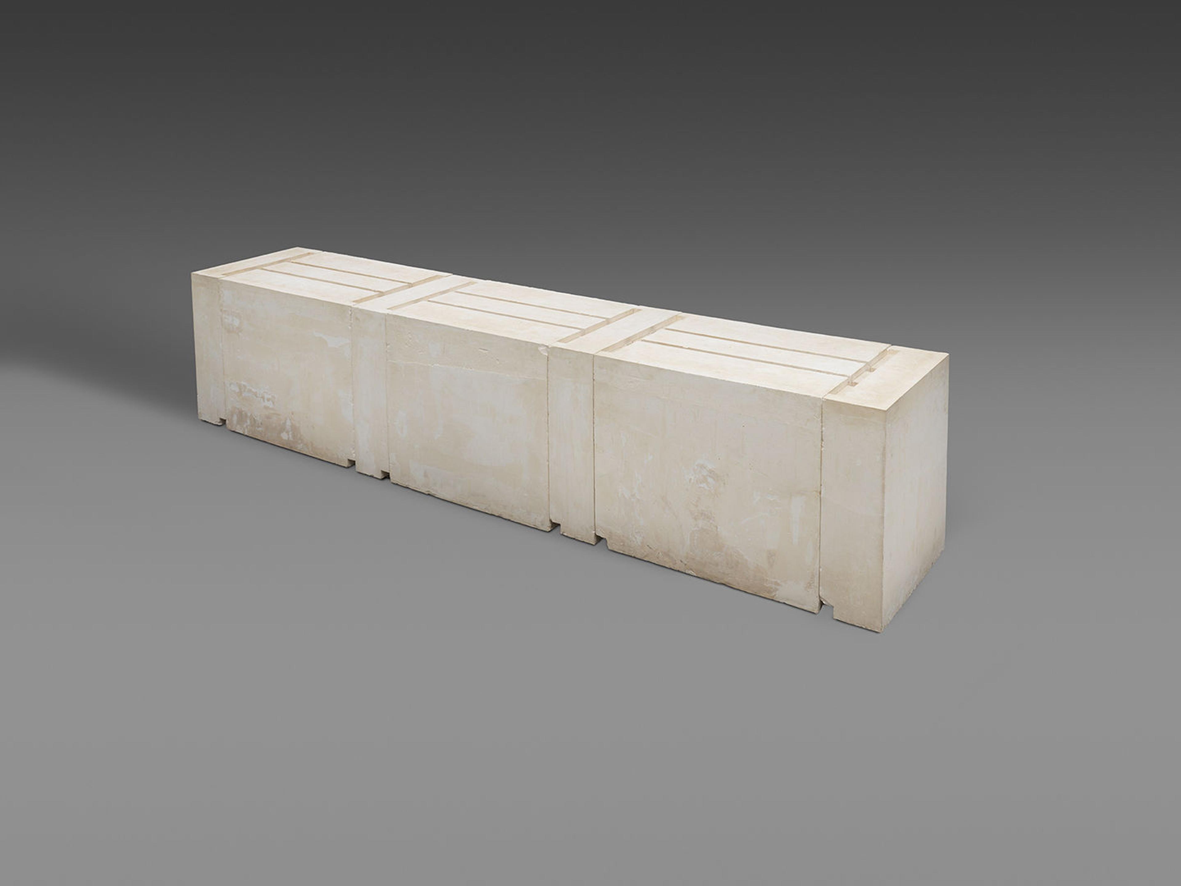 Bulky, rectangular plaster sculpture against a gray background. Grooves on the upper side of the sculpture outline the underside of three tables.