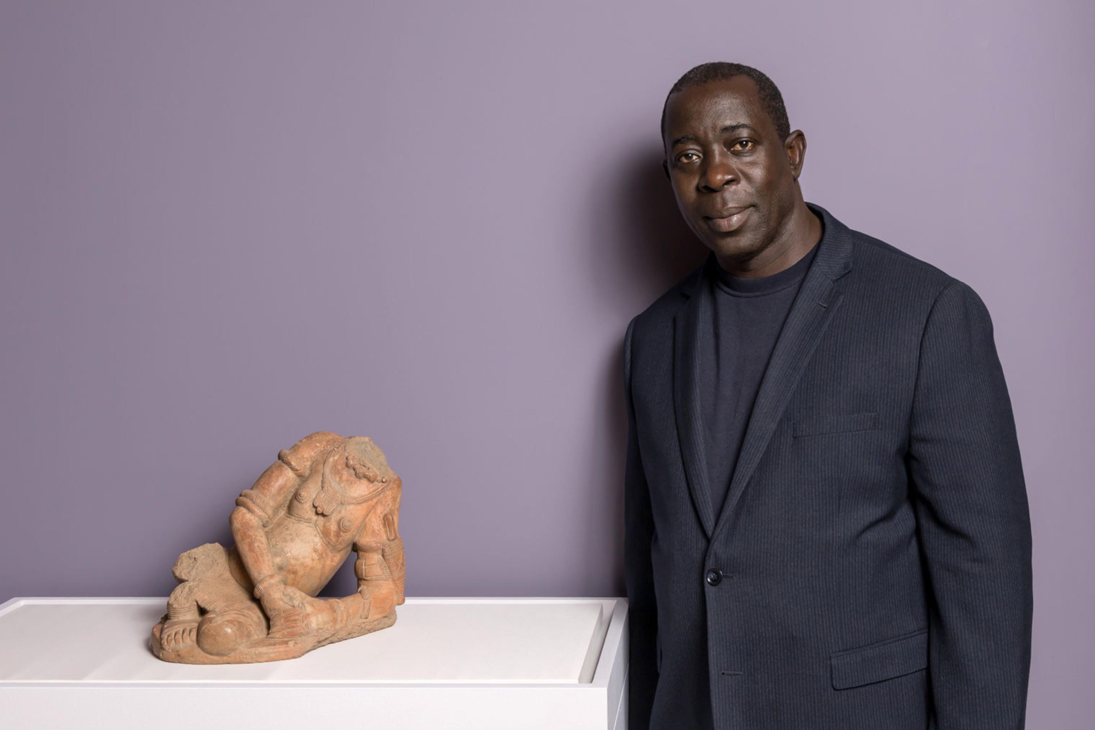 A tall man with dark skin, wearing a navy blue suit, stands next to a terracotta figure of a nude woman, in front of a purple background