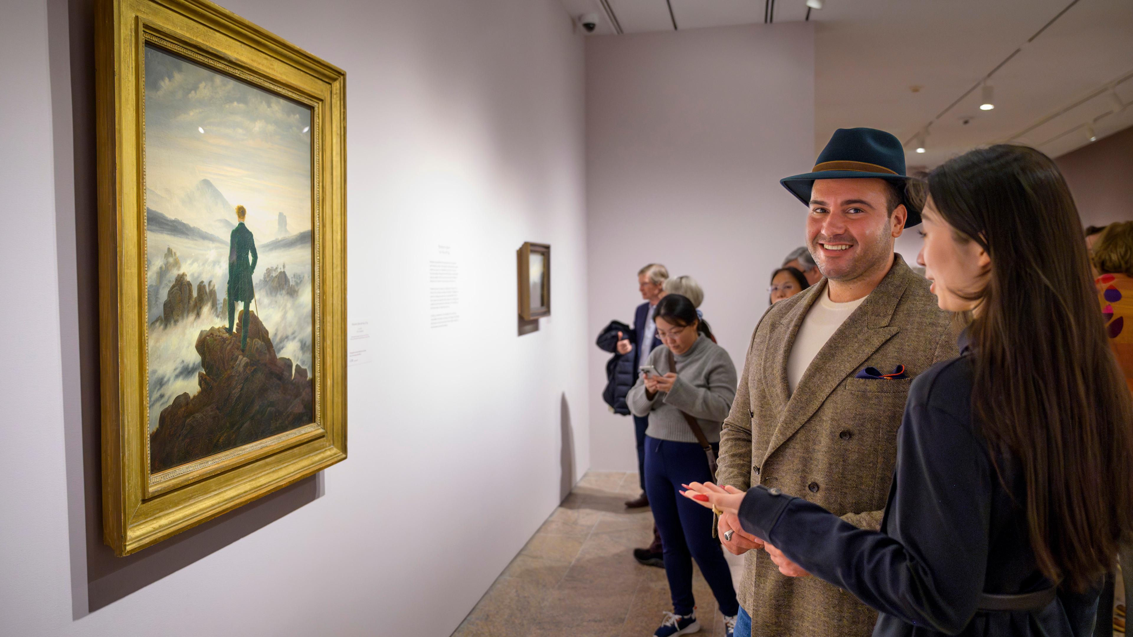 A man and woman stand in front of Caspar David Friedrich's painting "Wanderer Above the Sea of Fog" at an Evening Reception for Met Patrons.