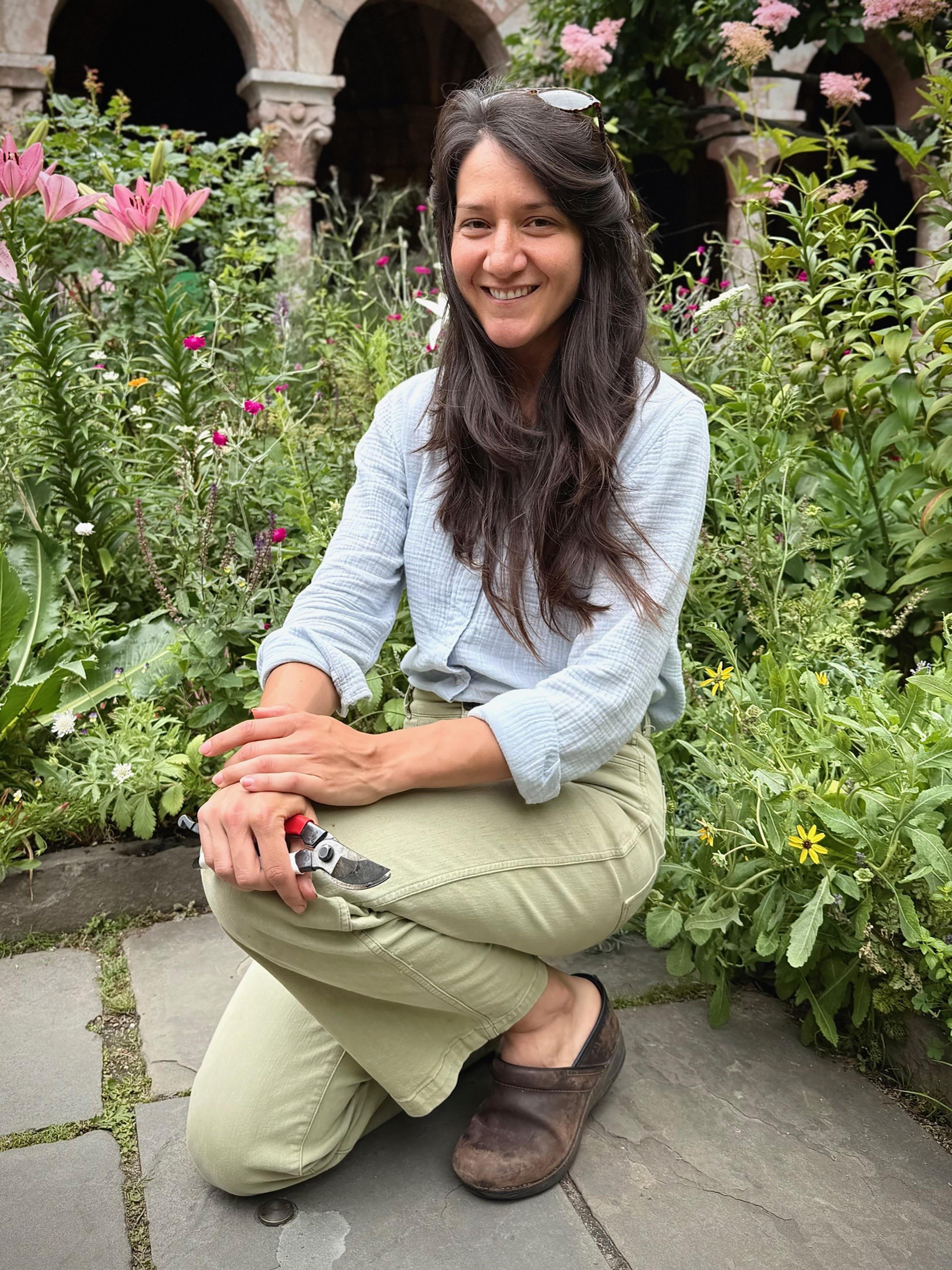 A woman with long brown hair kneels in a garden and smiles
