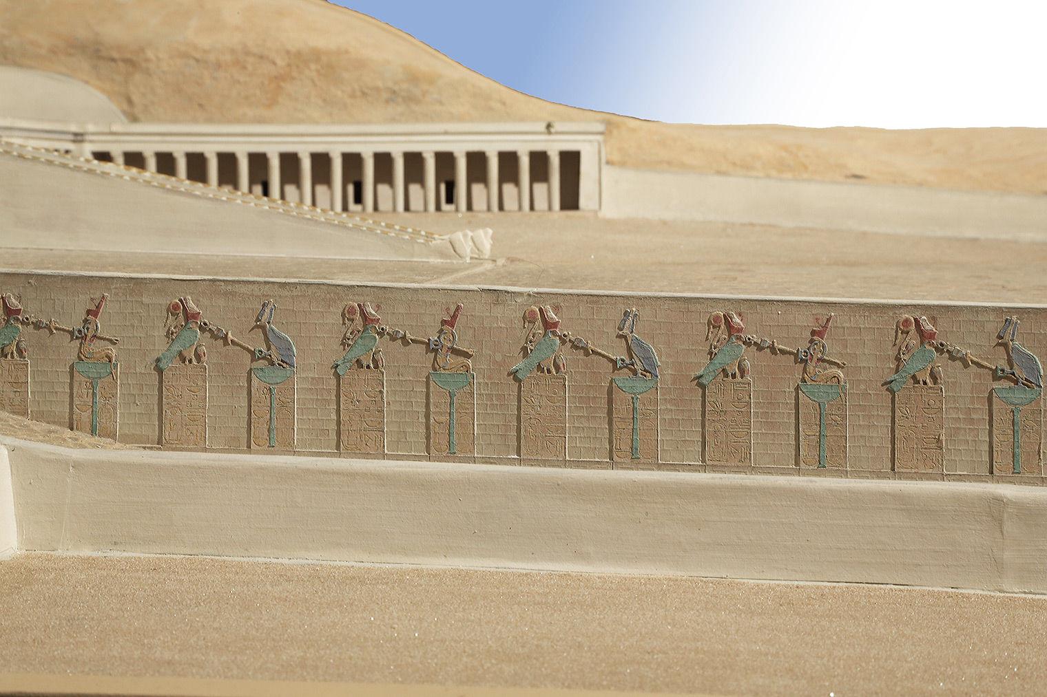 View of a stone wall from the side, with rectangular divisions topped by crowned falcons and cobras. The animals are green and the crowns red and white. A temple is in the background.