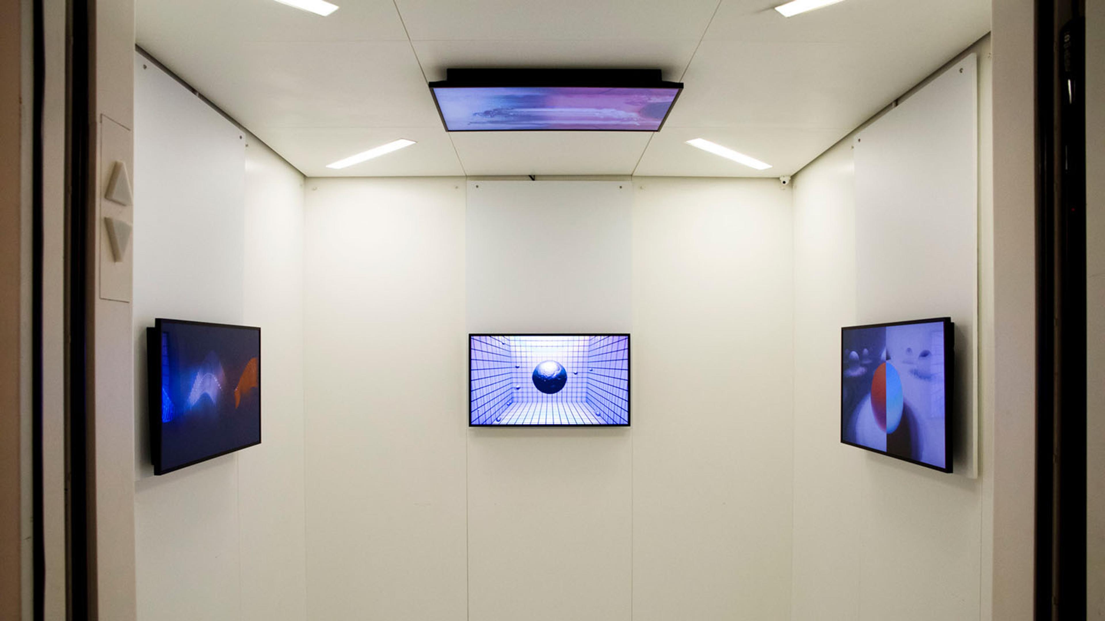 An elevator with the doors open and four video monitors displaying grids and geometric shapes hung on each of three walls and one mounted on the ceiling.