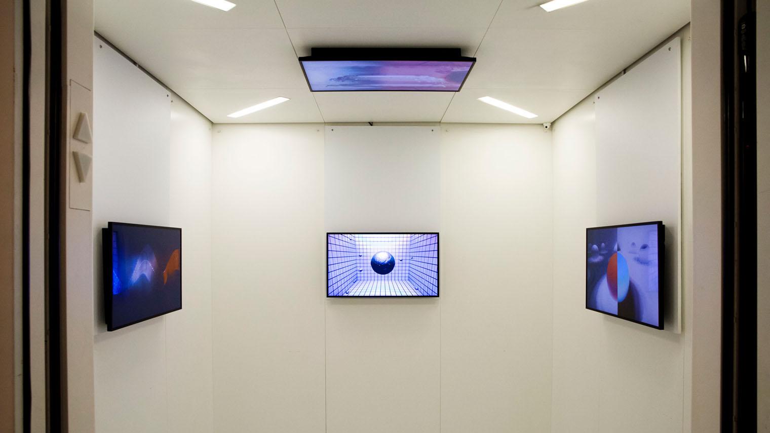 An elevator with the doors open and four video monitors displaying grids and geometric shapes hung on each of three walls and one mounted on the ceiling.