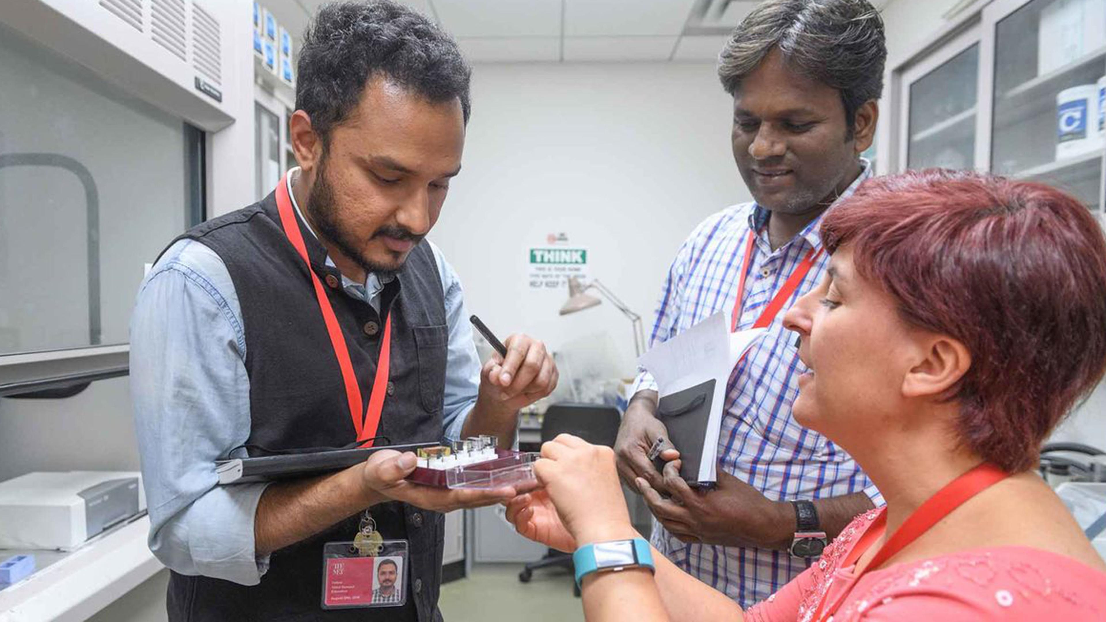 Three people in a lab collaborate on a project. One man holds a tablet with samples, while a woman points at it and another man observes. They wear ID badges and red lanyards, amid scientific equipment in the background.