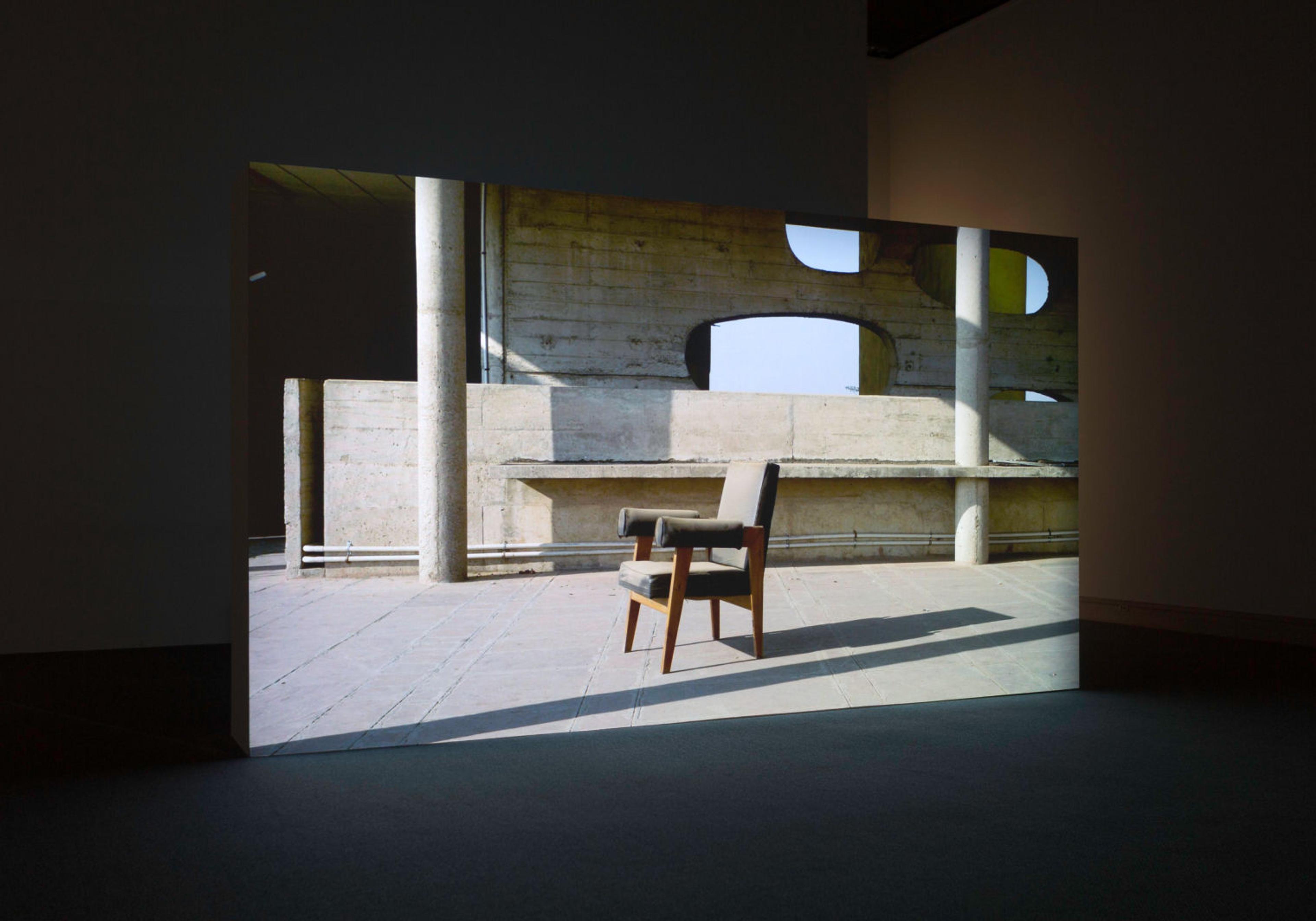 Gallery view of a screen showing a teak chair with gray cushions and armrests in an empty urban setting.