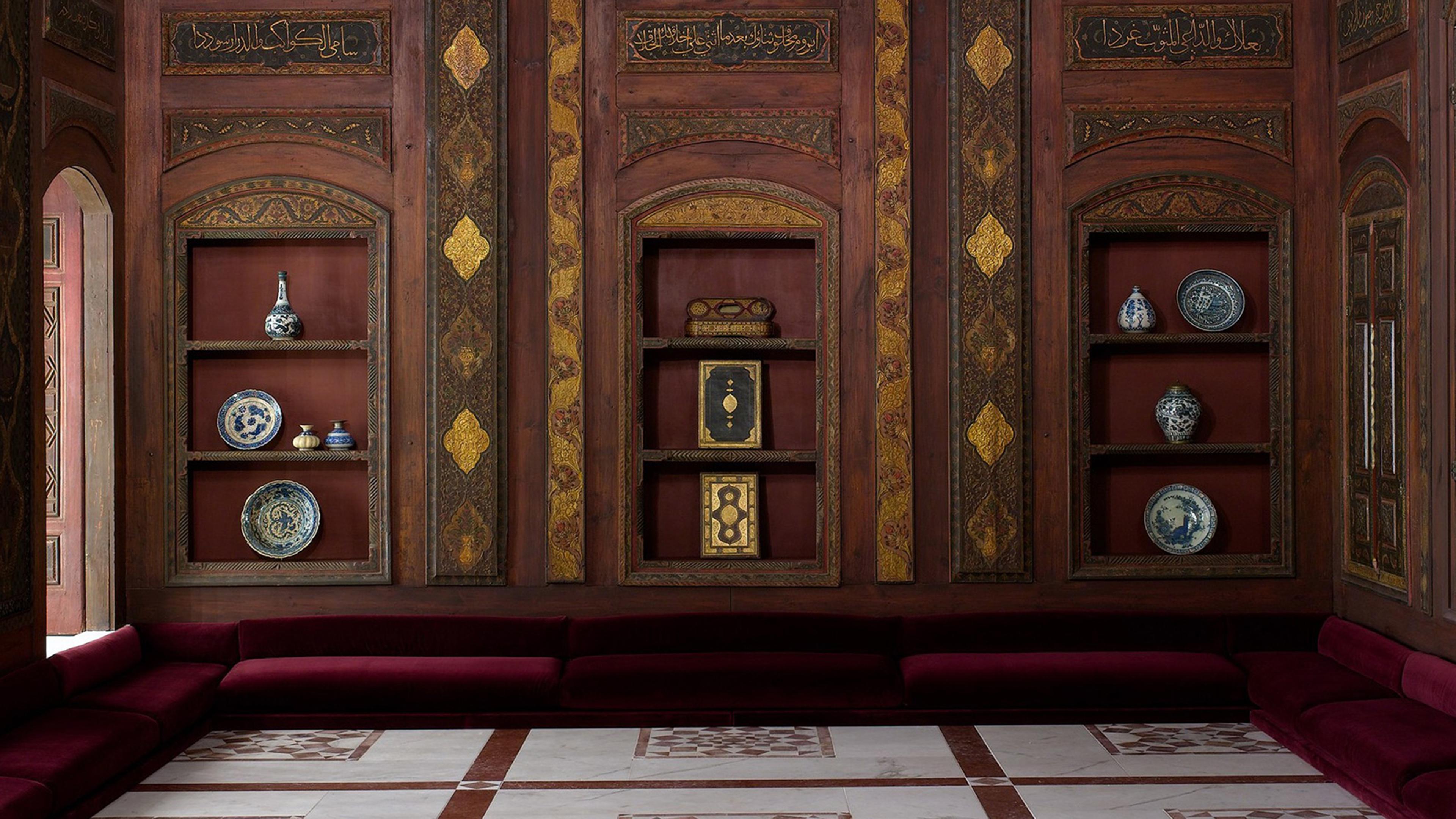 Photograph from a sumptuously decorated room with a geometric tiled floor, deep red seating cushions, and wooden wall panels detailed in gold leaf.