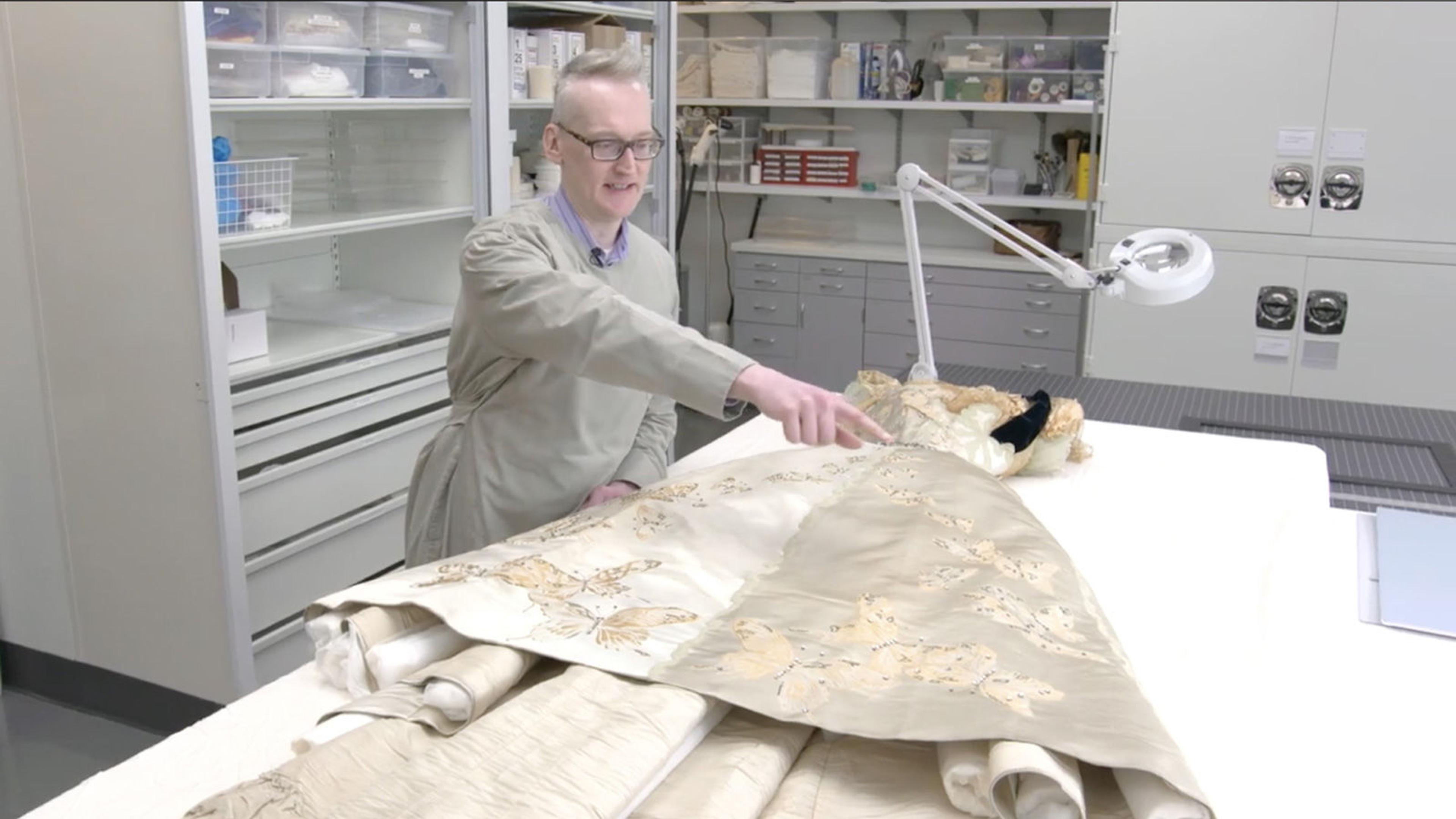 A seated conservationist points to an area on a beige ball gown, spread out on a white table in front of him.