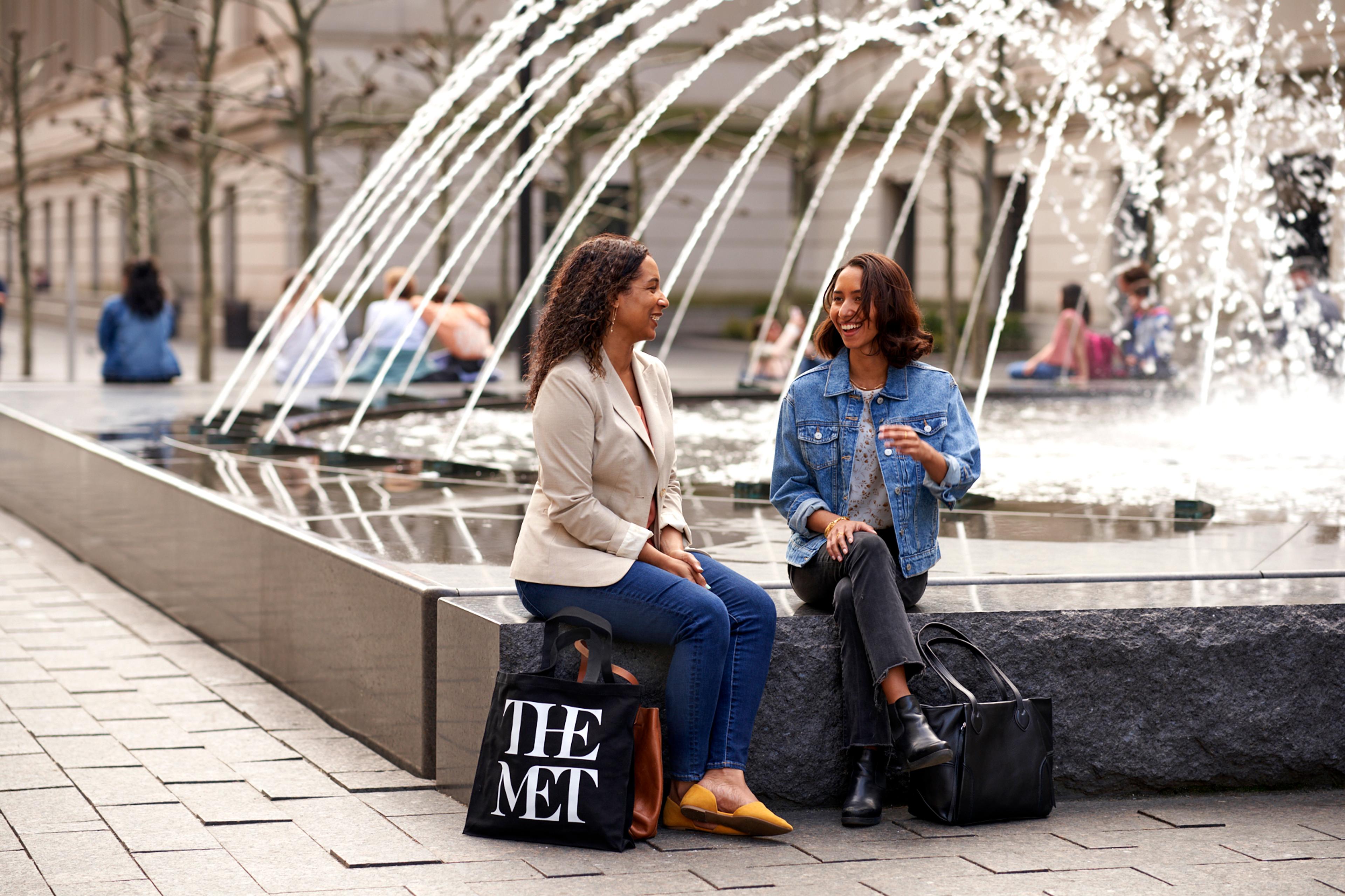 Two medium-skin-toned women sit by the fountains chatting on The Met Plaza.