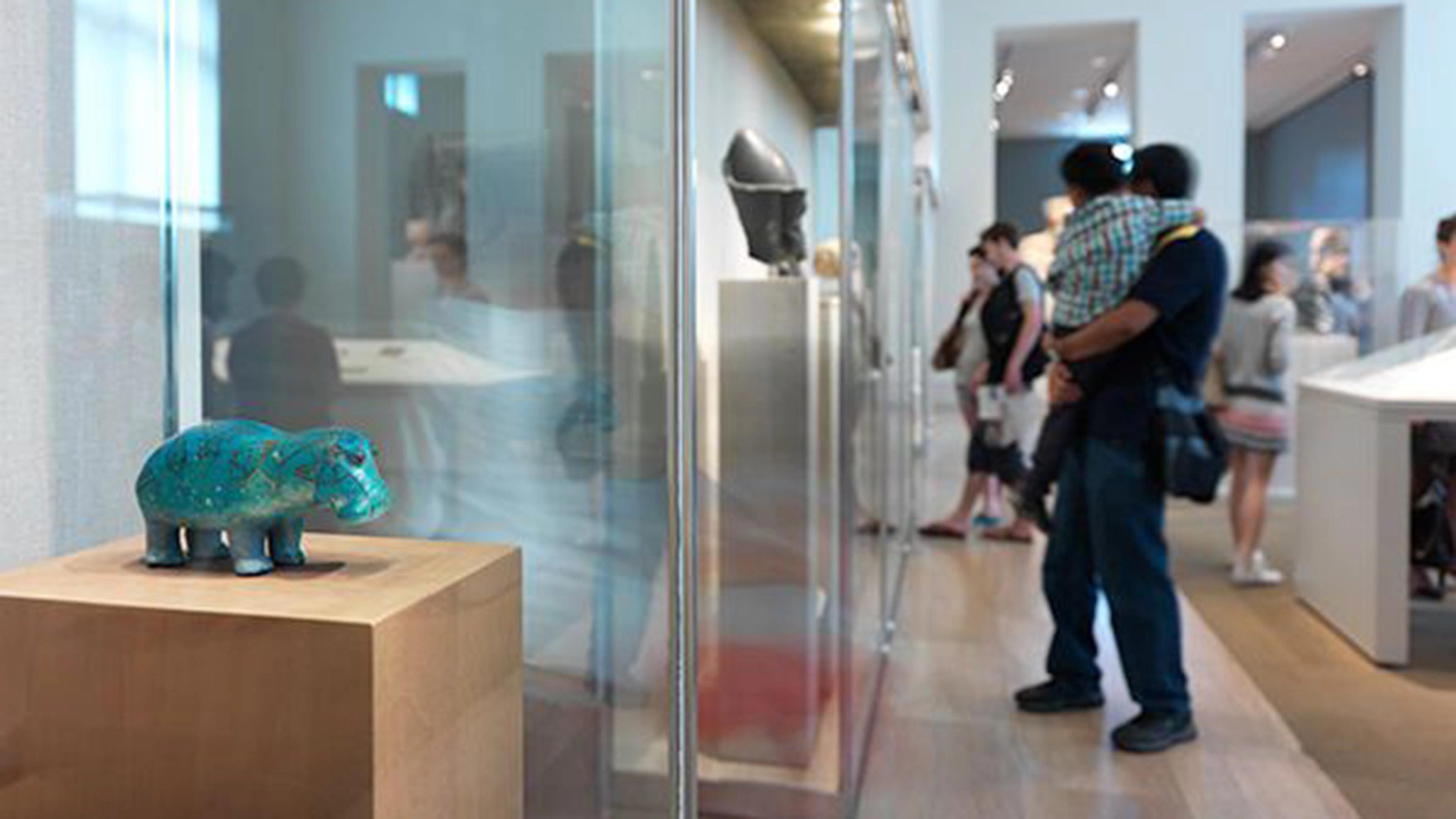 A teal ceramic hippo sculpture is displayed on a wooden pedestal in a bright museum, surrounded by visitors.