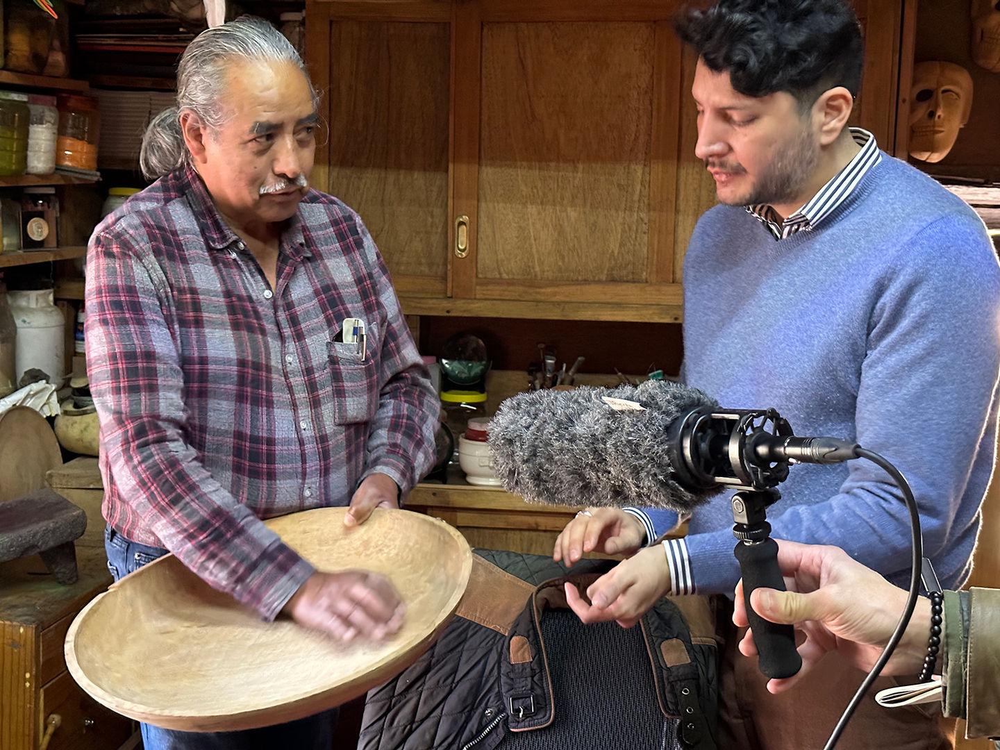 An older, casually-dressed man demonstrates a movement inside the bowl of a batea to a well-dressed man. A microphone is held in the center of the image by someone outside the frame.