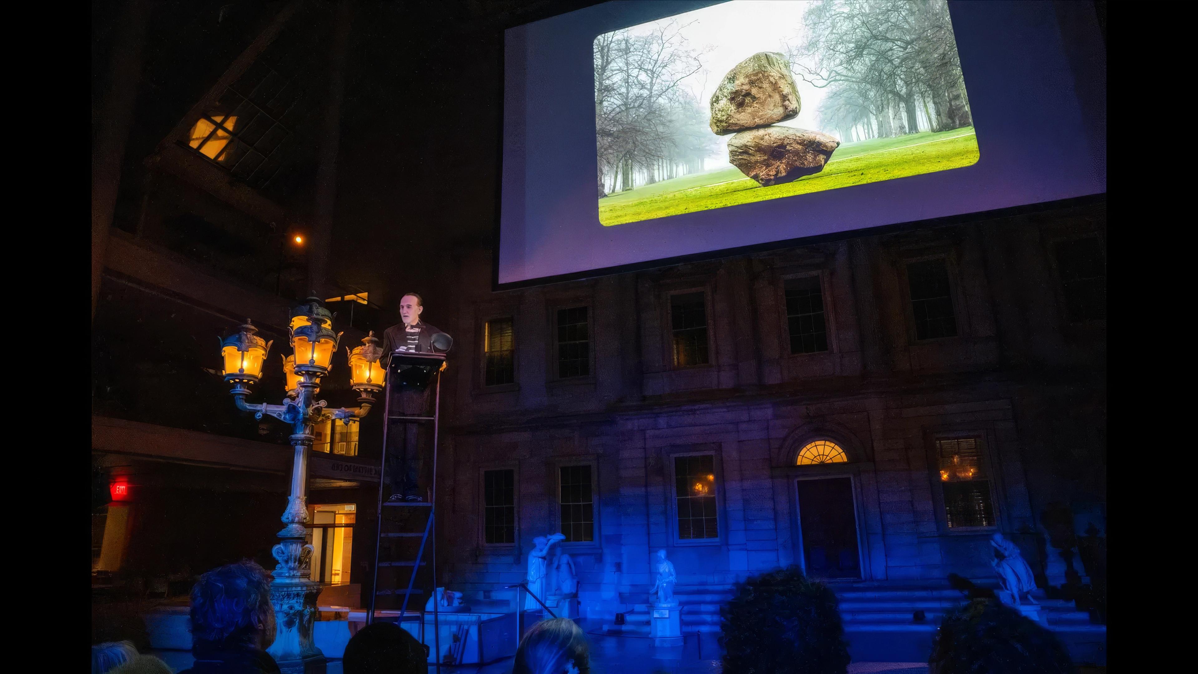 A person stands at a podium in a dimly lit gallery beneath a large projected image of two stacked rocks in a landscape.