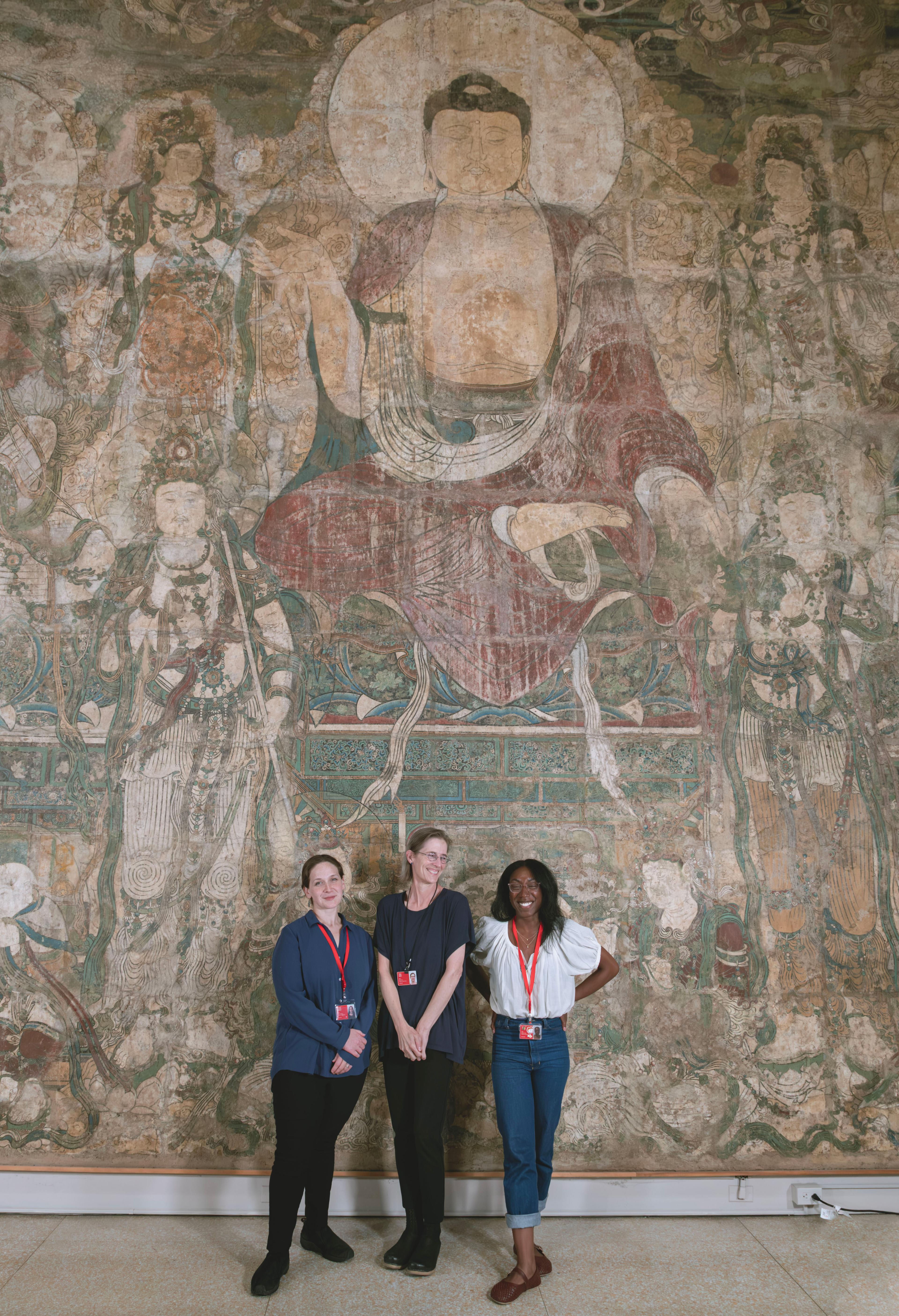 Three smiling Met staff members standing in front of a large, faded mural of Buddha.