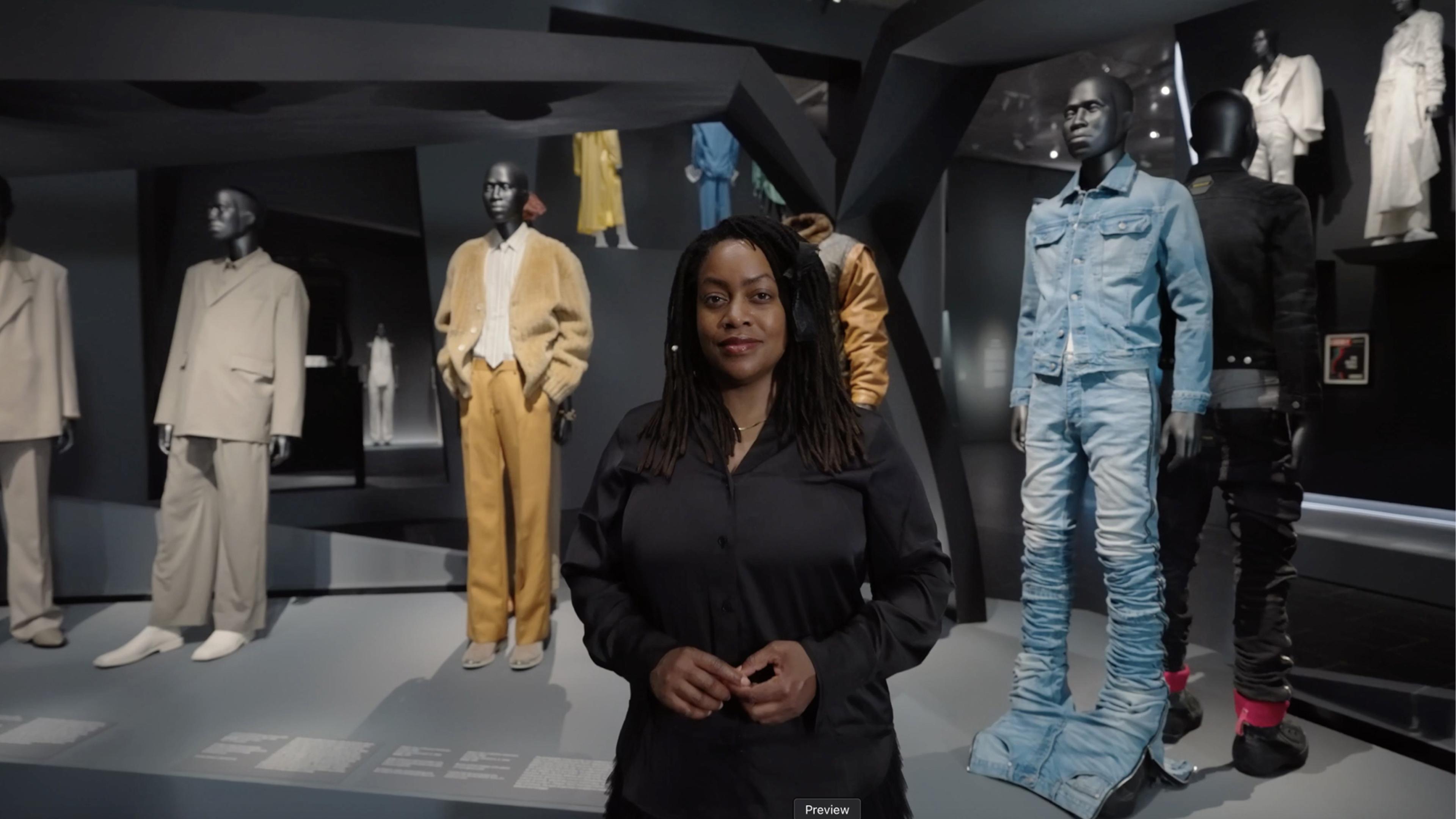 Black woman wearing all black, standing in front of mannequins dressed in blue, yellow and beige.