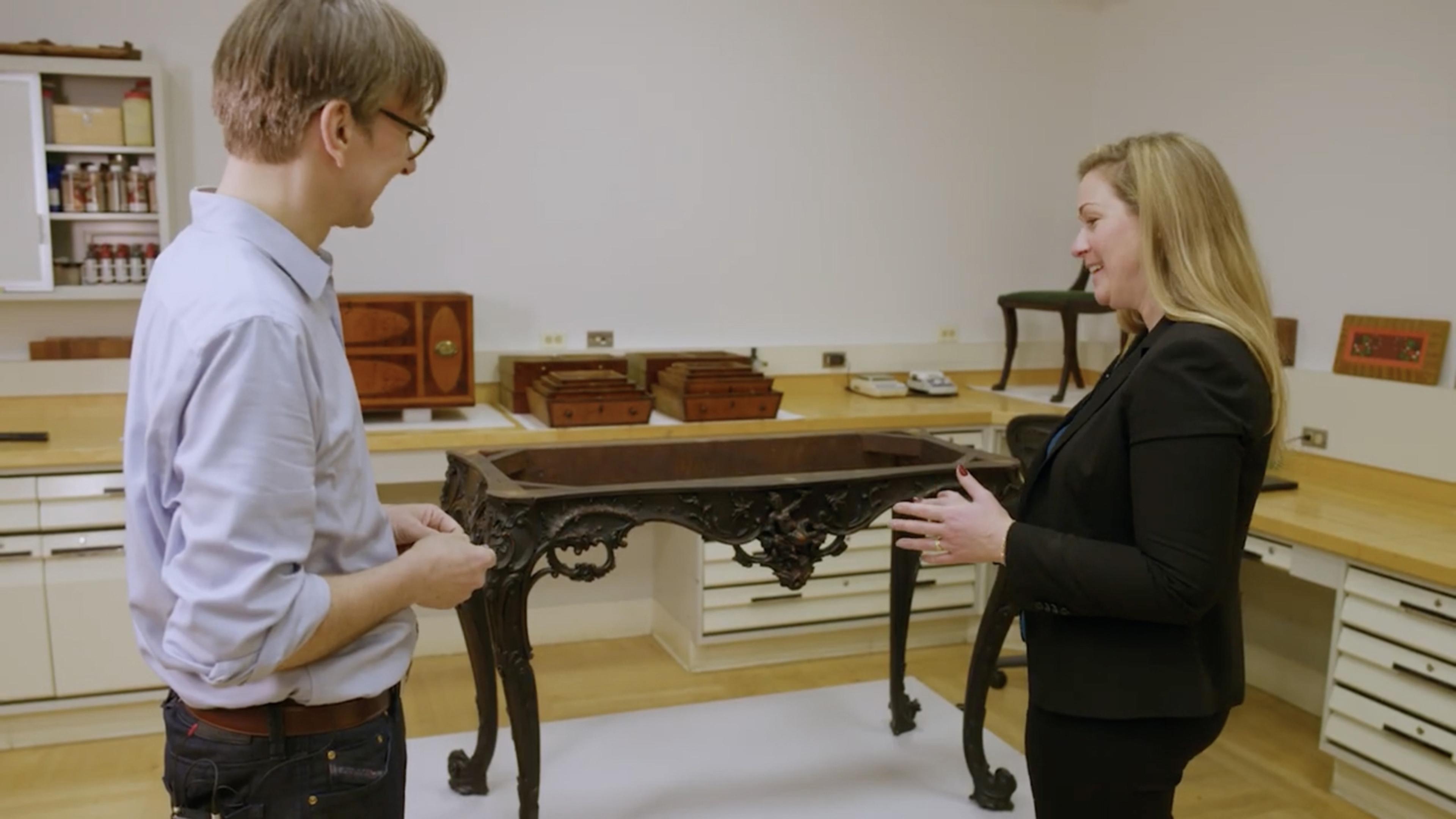 Two people stand in front of an ornate table in a workshop space. The table has curved legs and elegant detailing, and is missing its top.