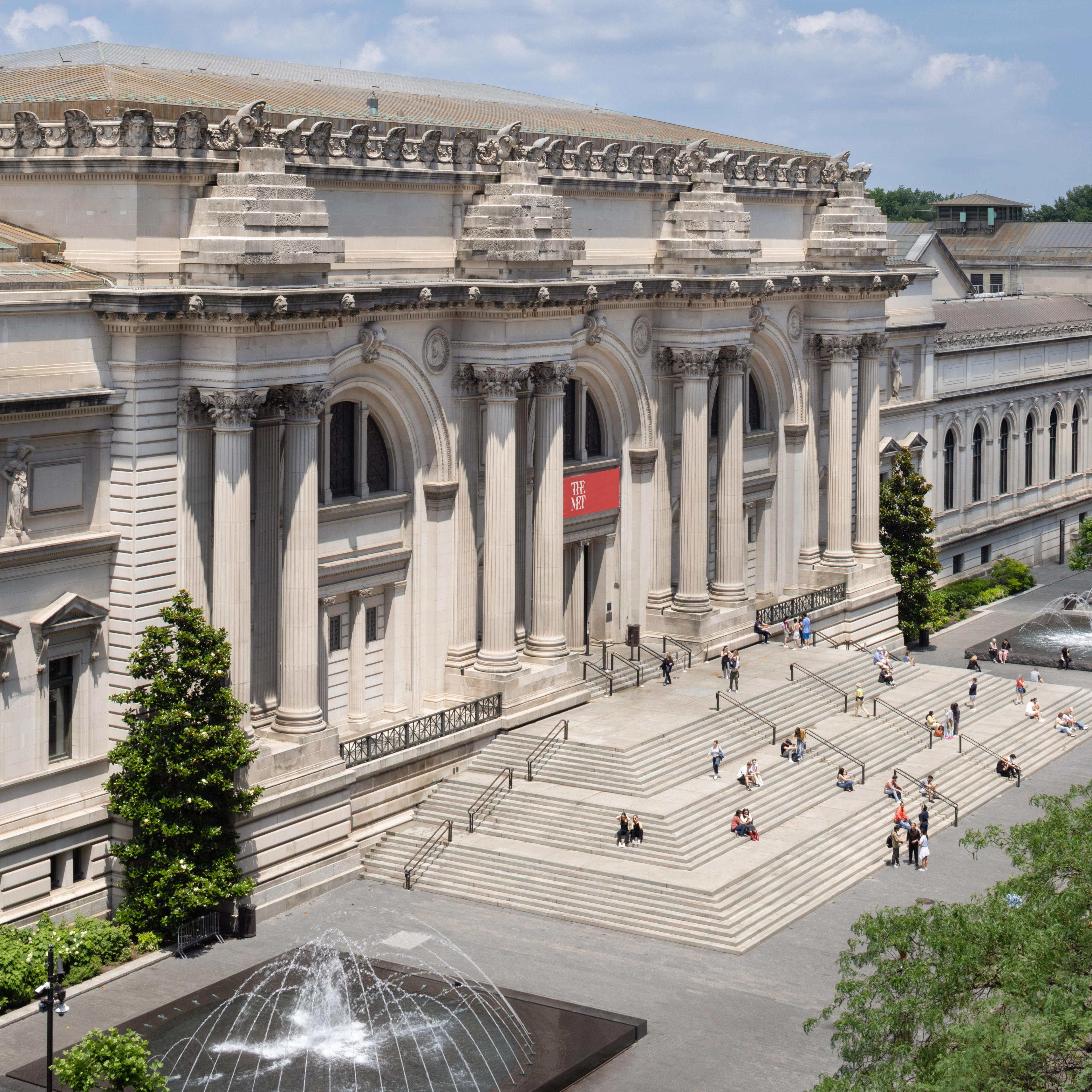 The front of the Metropolitan Museum of Art building with steps, people walking, a fountain, and yellow taxis parked along the street.