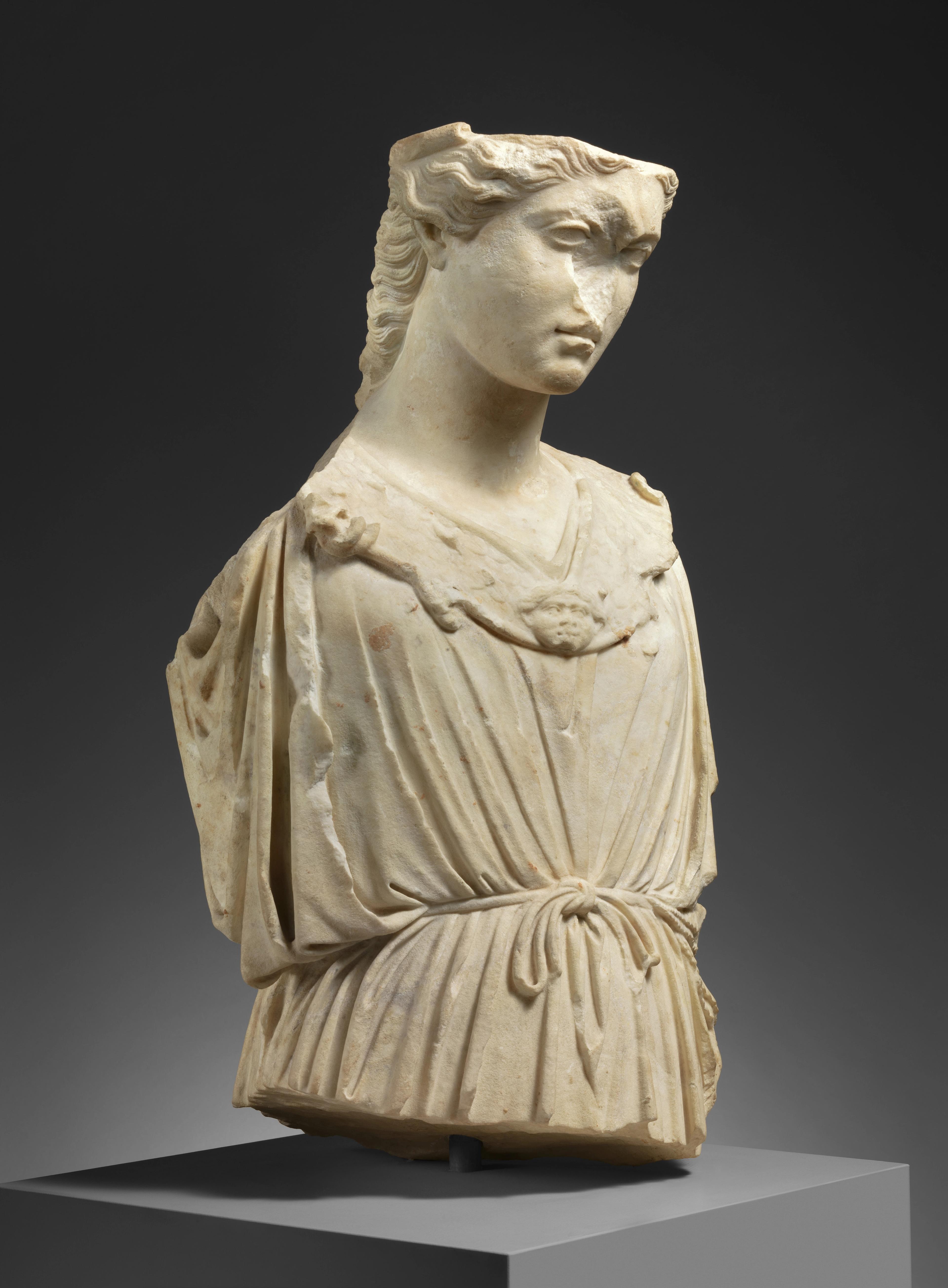 Marble bust of Athena with her head angled downwards. Her features appear somber; the sculpture shows signs of wear and is missing a nose.