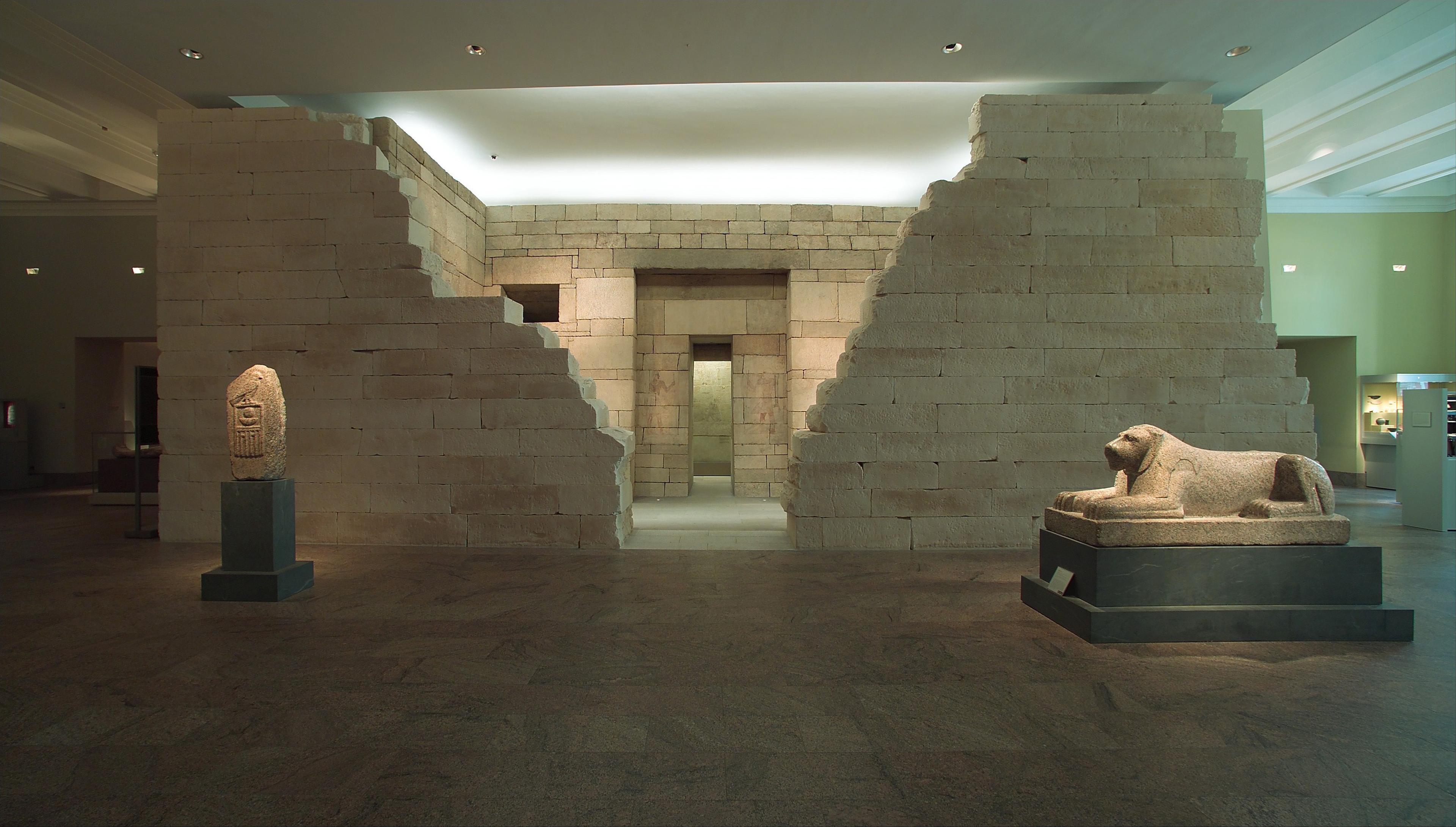 Museum gallery with a stone temple featuring a central doorway and sloping walls, flanked by a carved standing figure on the left and a reclining sphinx sculpture on the right.