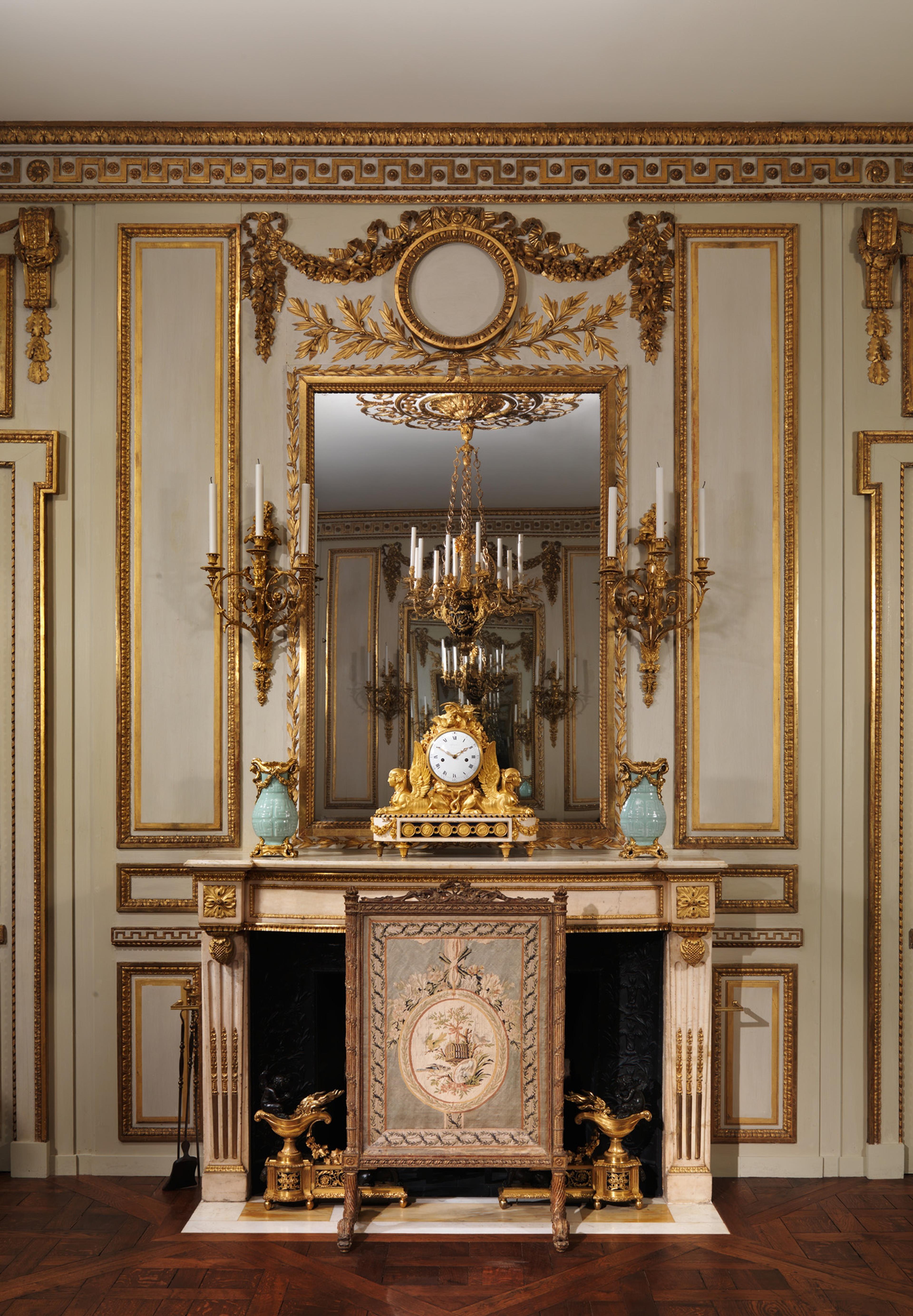 A richly decorated room with ornate gold-trimmed wall panels, a marble fireplace topped by an elaborate gilded clock and vases, and a large mirror reflecting a chandelier.