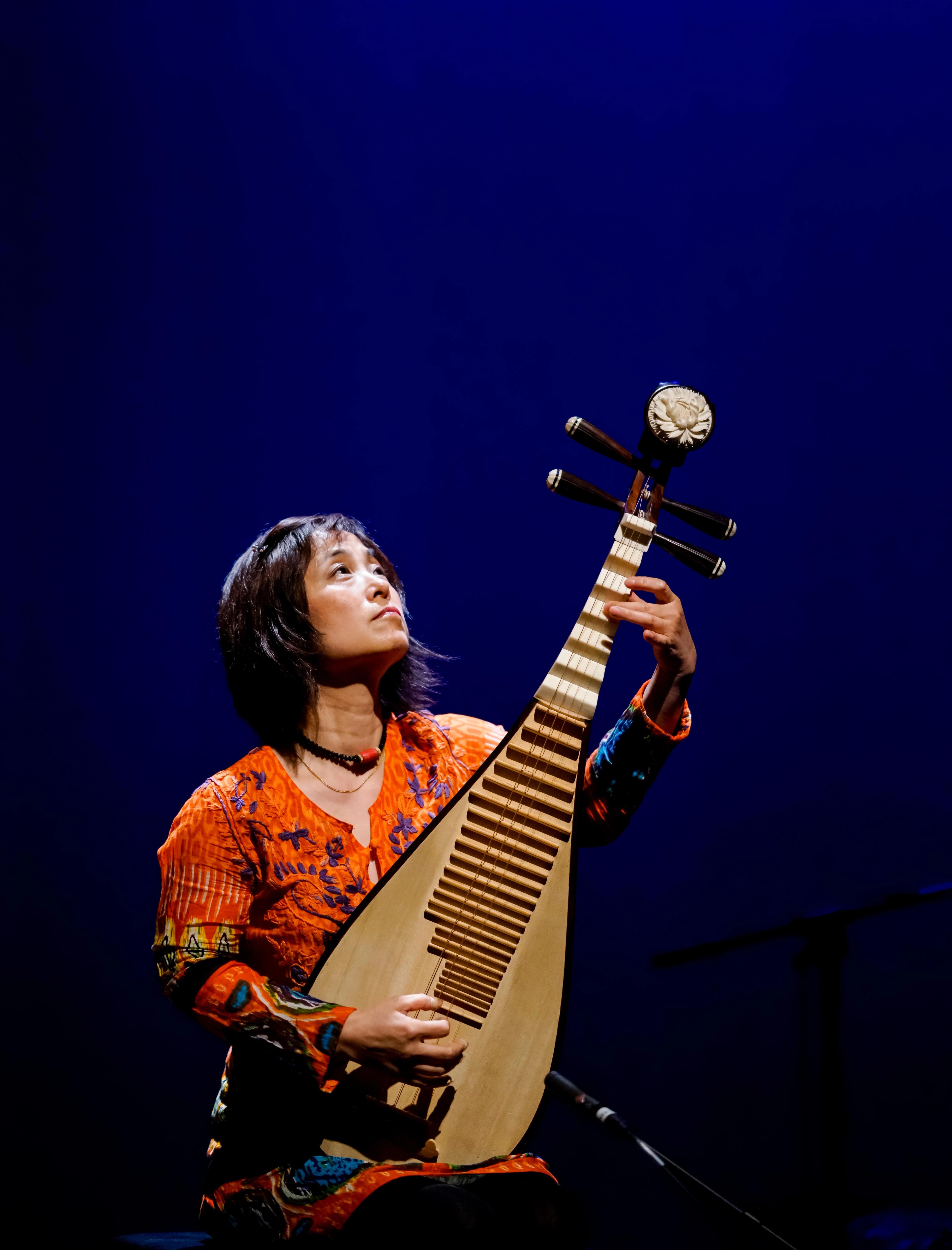 Wu Man holding the pipa and looking upwards against a dramatic, deep blue background.