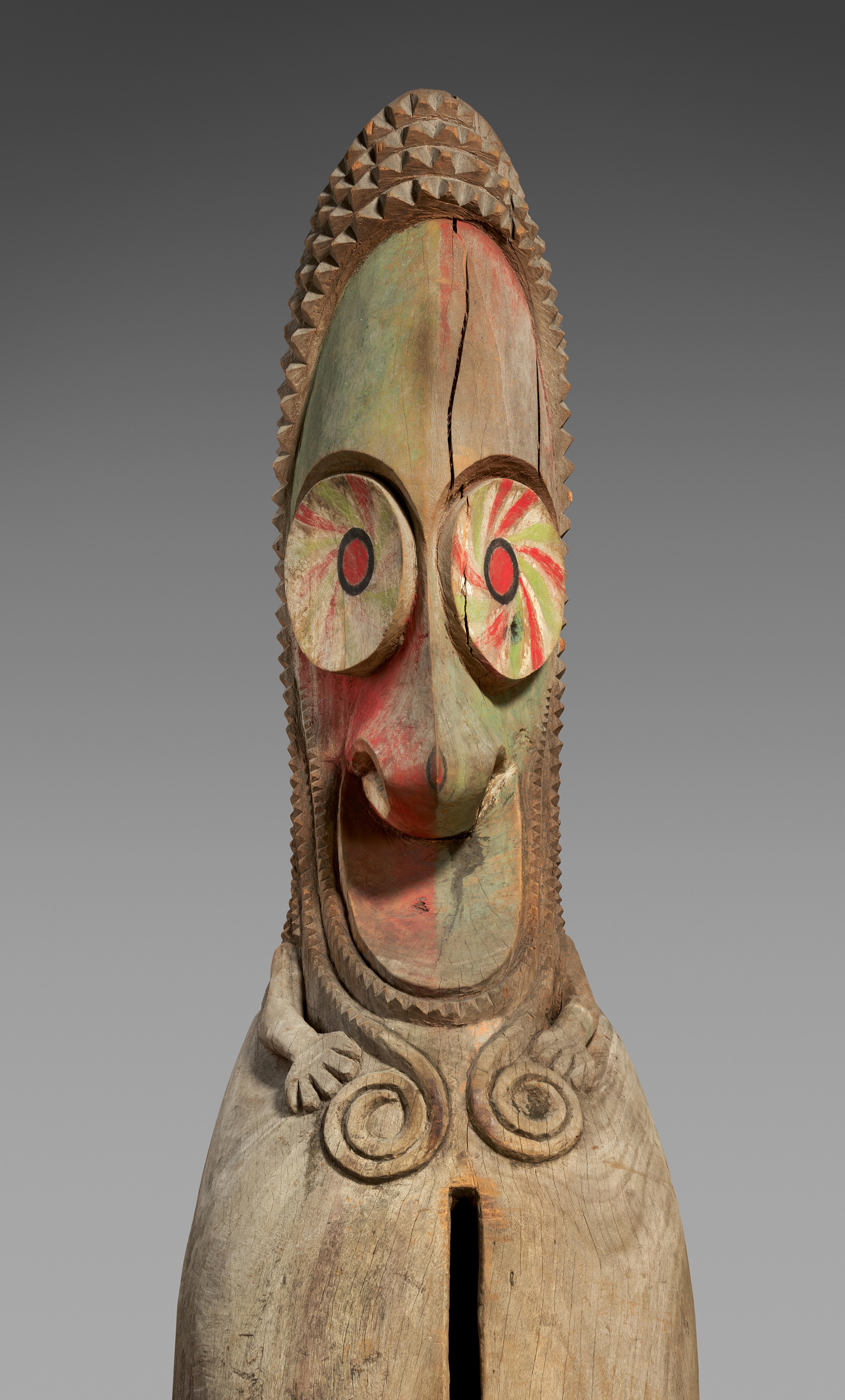 Carved wooden figure with an elongated face, large circular eyes painted with red and green spirals, and a wide open mouth.