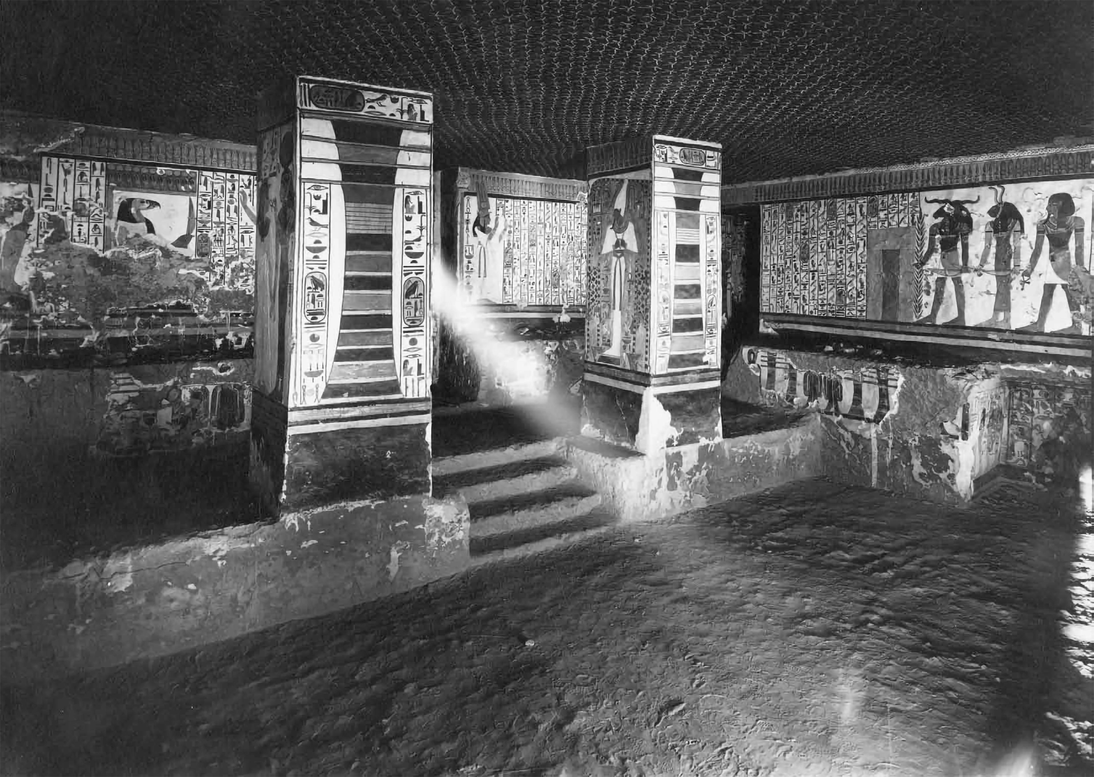 Black-and-white photograph from the inside of an Egyptian tomb. Hieroglyphics and scenes are painted on the tomb's walls. A beam of light shines between two pillars.