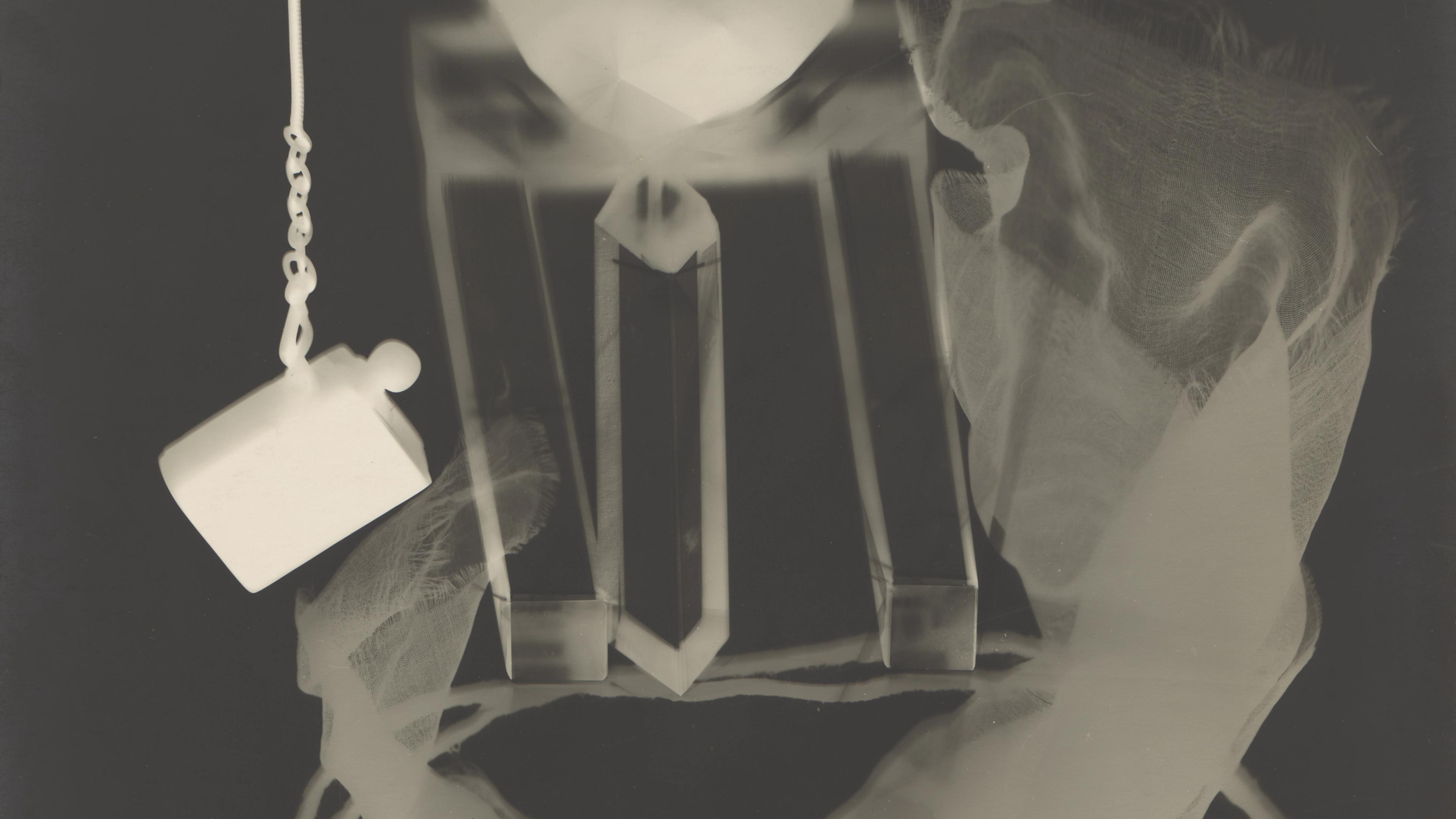 A black-and-white abstract photogram of overlapping translucent forms, including a prism and a small hanging object.