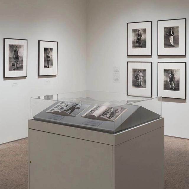 A gallery room with black-and-white portrait photographs on the walls and a display case containing open books in the center.