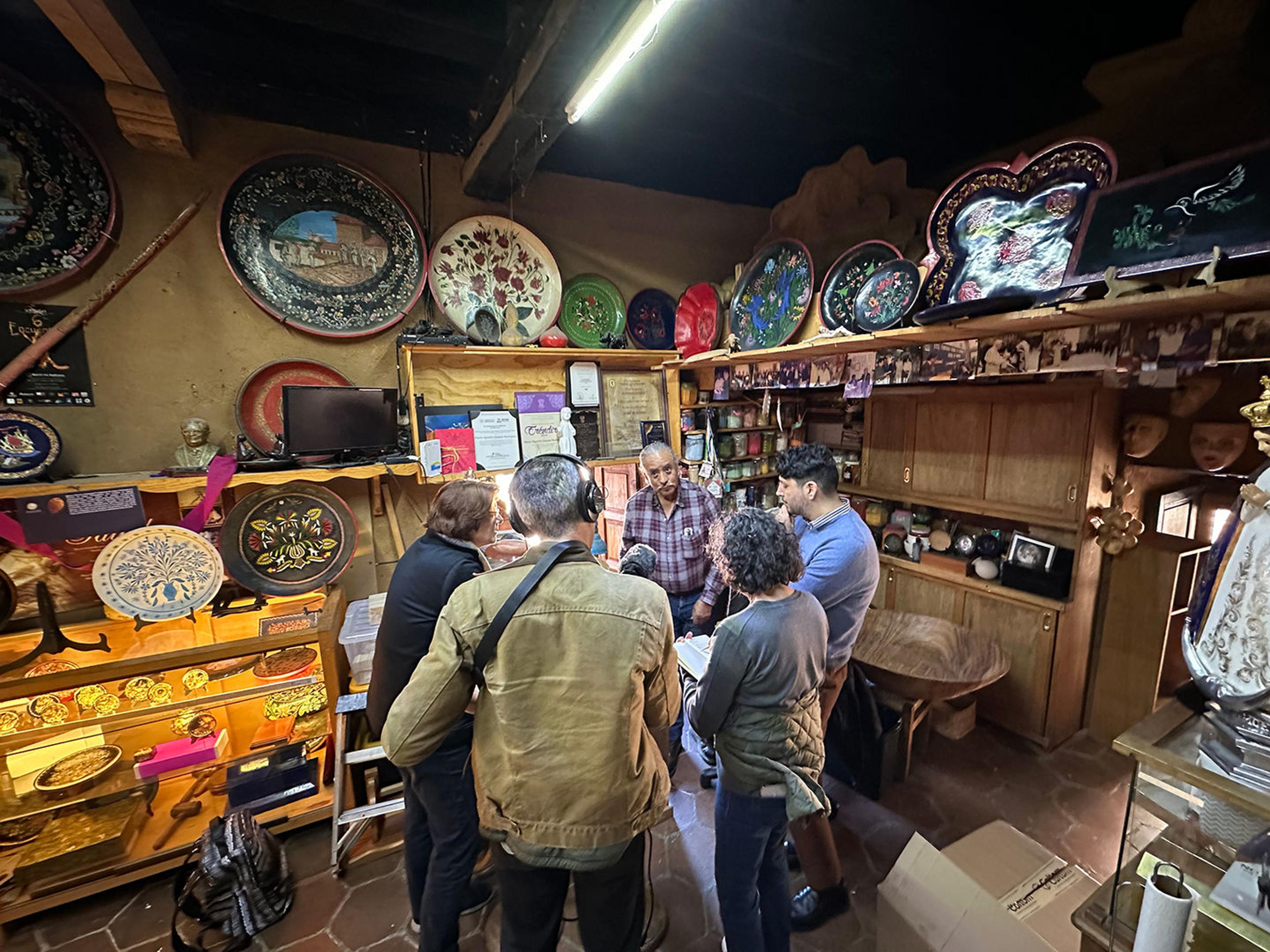 Photo in a workshop with numerous decorative trays on display. Four people listen to a man speaking in the back corner of the workshop.