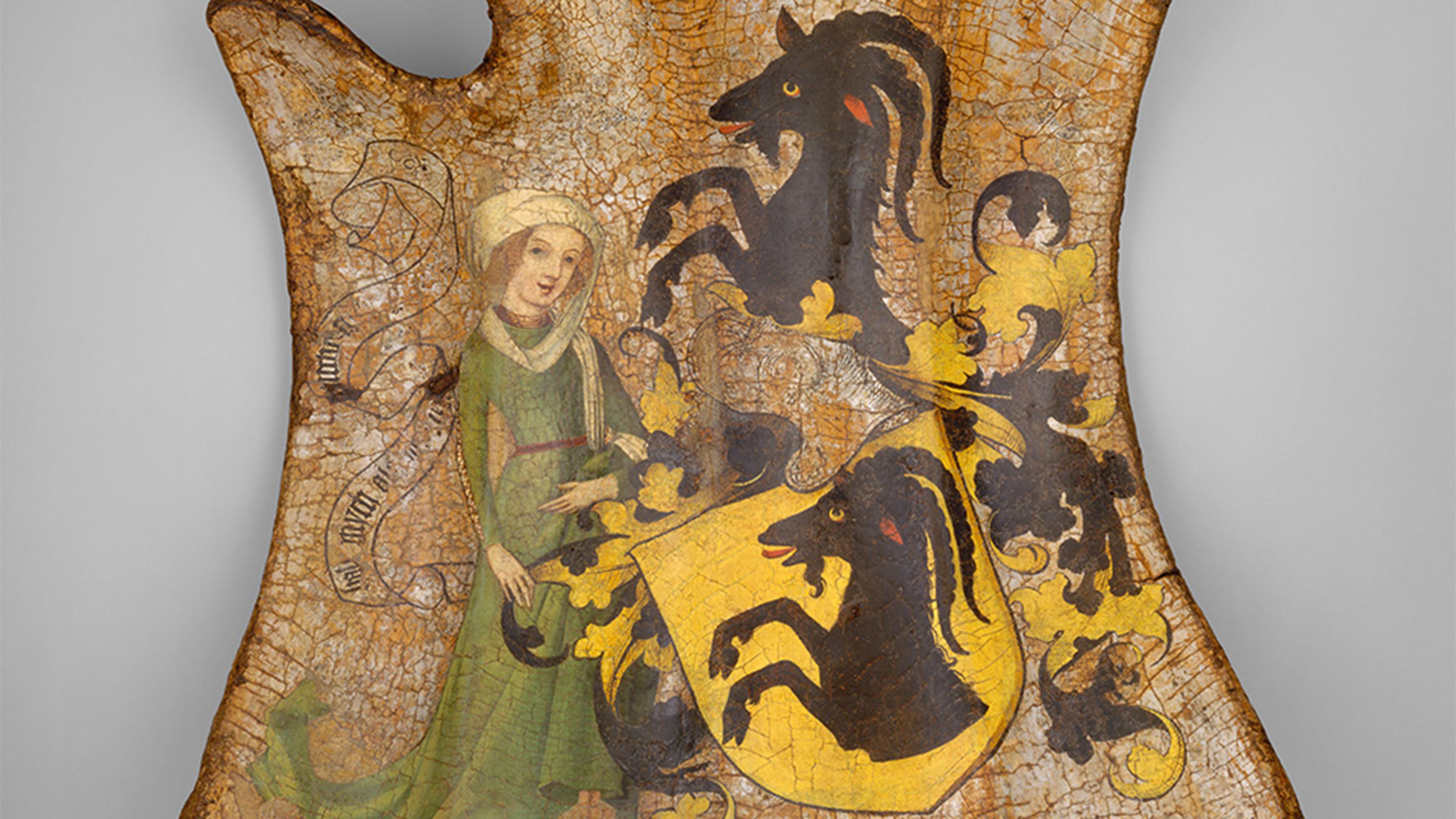A ribbed shield painted with a woman in green beside a black goat on its hind legs and a yellow crest also depicting a goat.