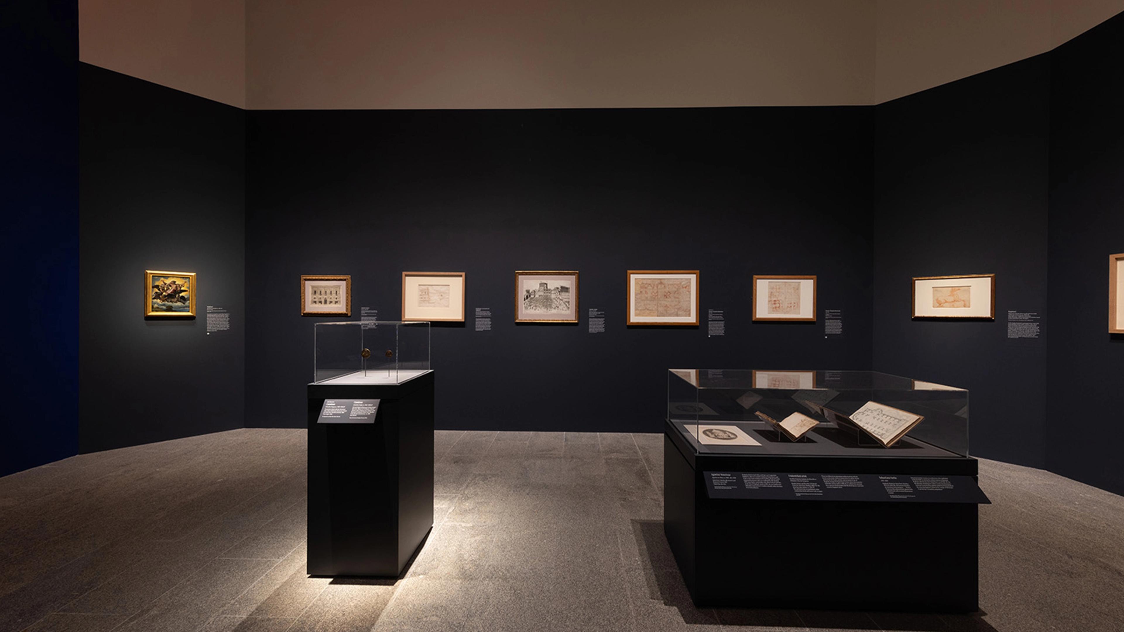 A dimly lit museum gallery displays framed artworks on dark walls and books in glass cases.