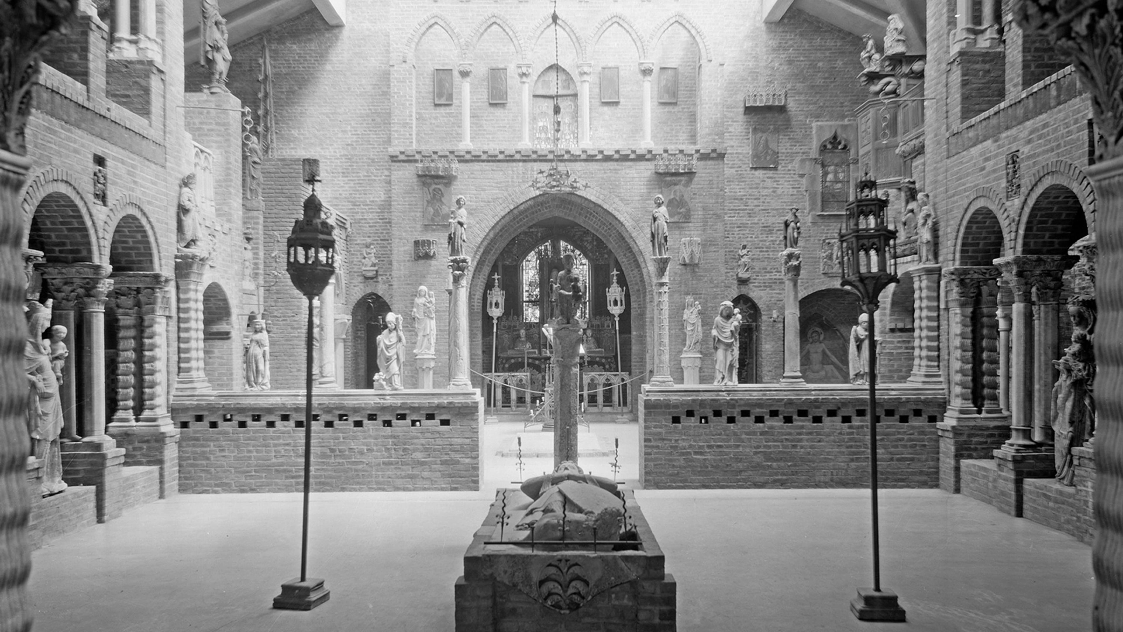 Medieval-style hall with brick arches and statues, illuminated by lanterns arranged around a central stone structure.