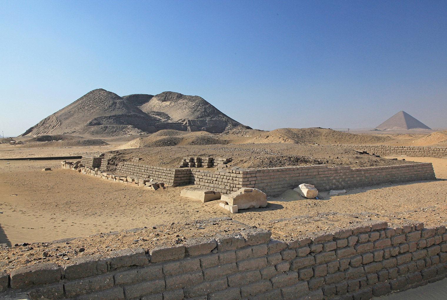 Mudbrick remains of wall in the foreground, remains of a mudbrick structure behind it, and in the background a decayed pyramid to the left and a preserved pyramid to the right
