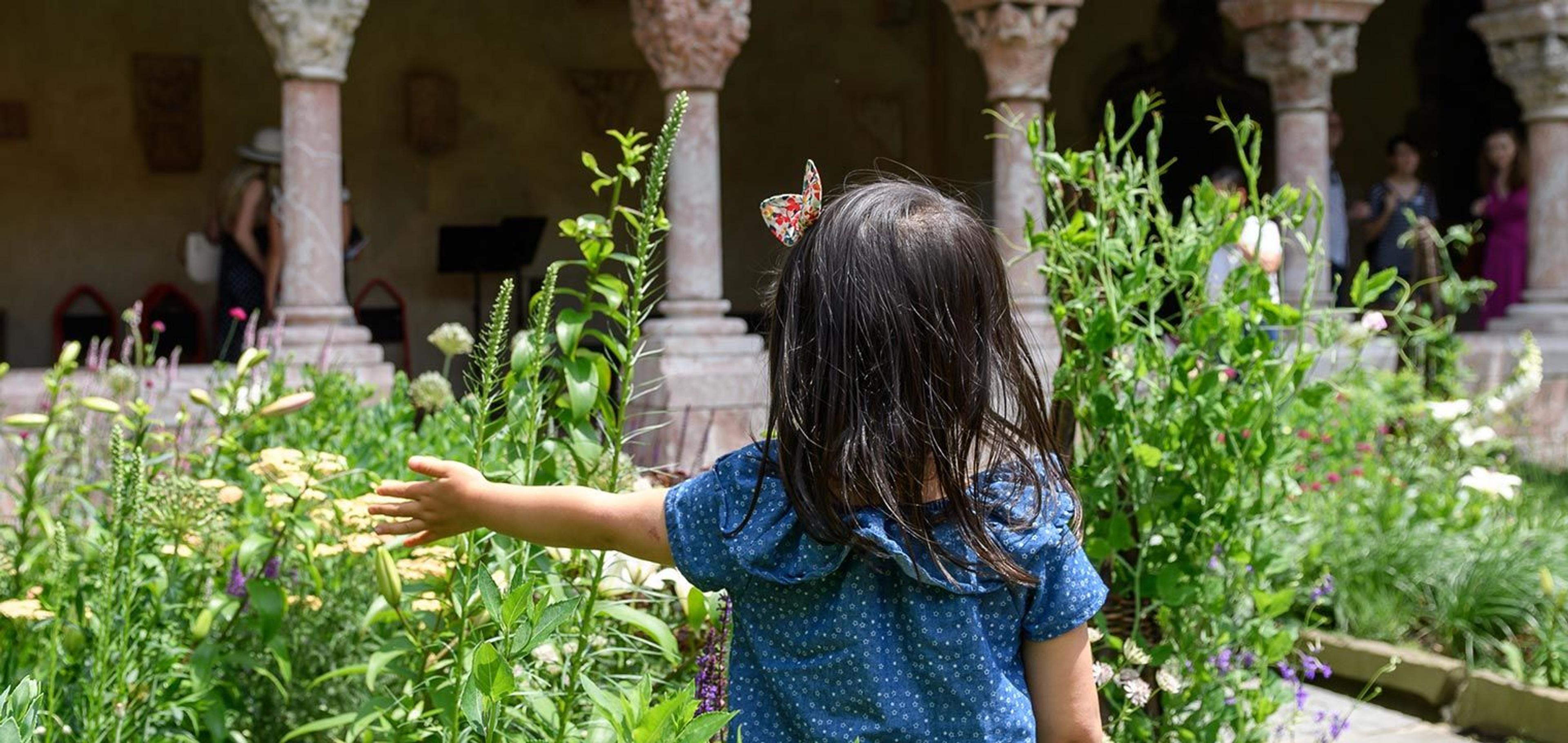 a young girl with her back turned to the camera stands in front of a garden bed in a medieval cloister.