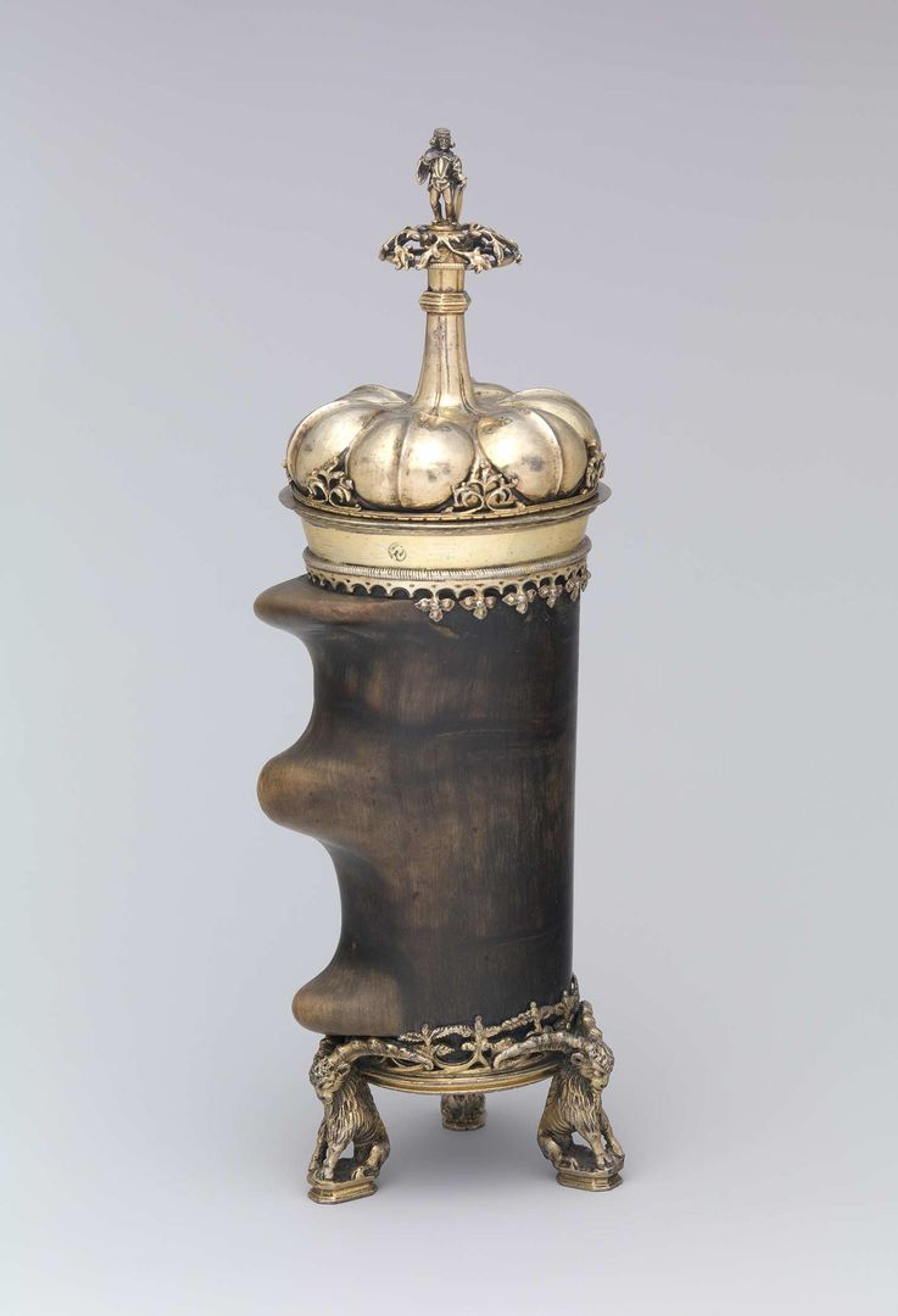 This drinking vessel has three metal legs and an ornate metal lid that rises into a small platform with a figure of a nobleman on the top. The body of the vessel is a dark brown section of a mountain goat horn with two ridges on the left side.