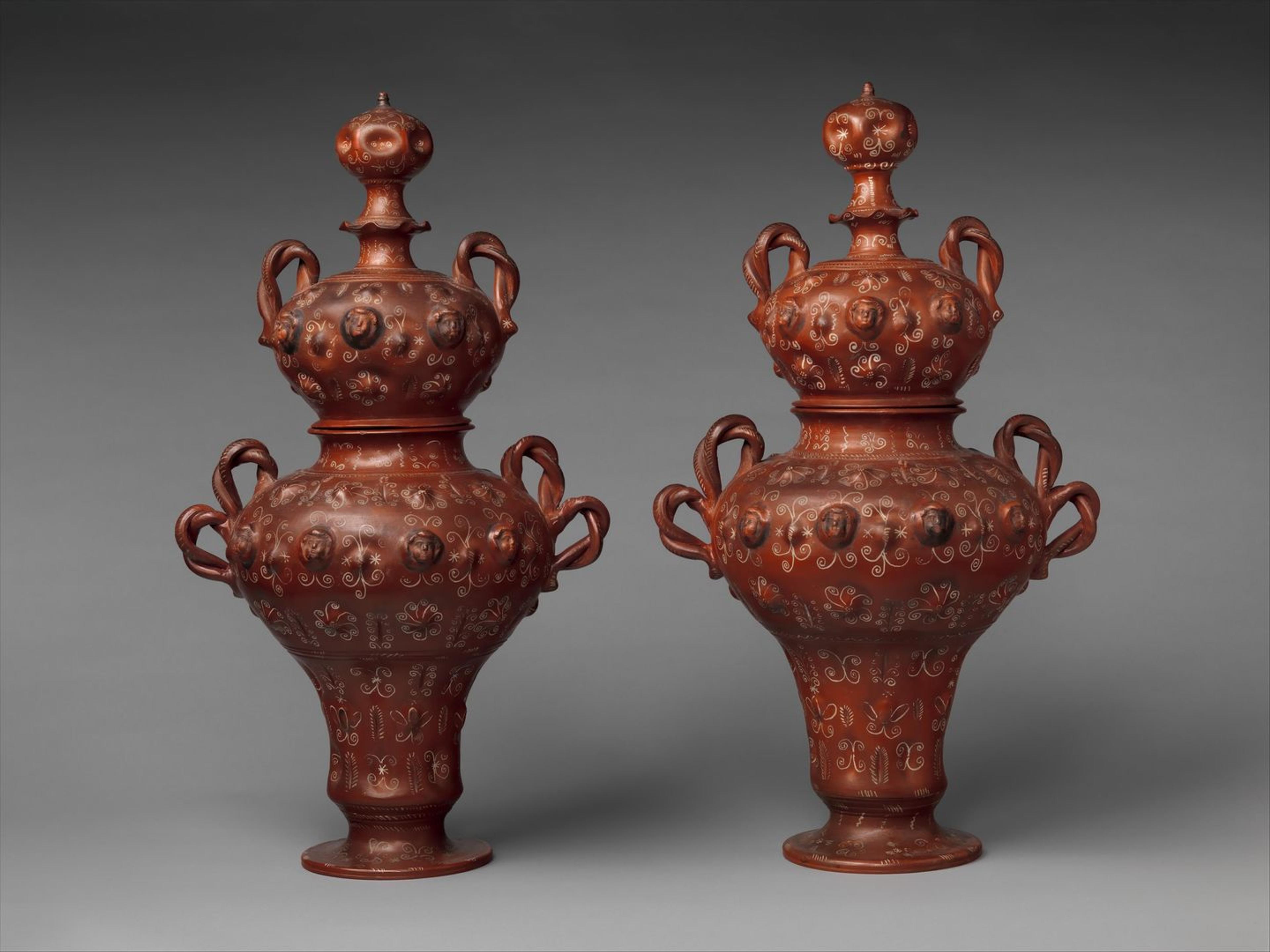 Photograph of two red colored jars with white patterns and sculpted faces on top, against a grey backdrop.