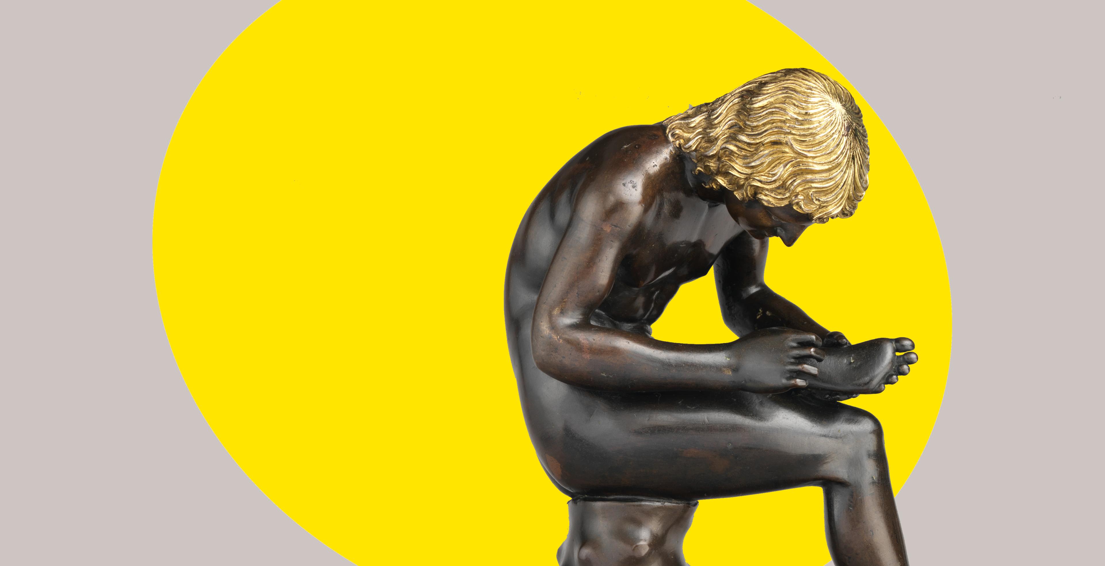 A small bronze sculpture of a seated boy pulling a thorn from the bottom of his foot appears within a display case at The Metropolitan Museum of Art; the figure hunches over looking intently at the bottom of his foot; the sculpture is set against the warm walls and limestone columns of the against gallery in the distance.