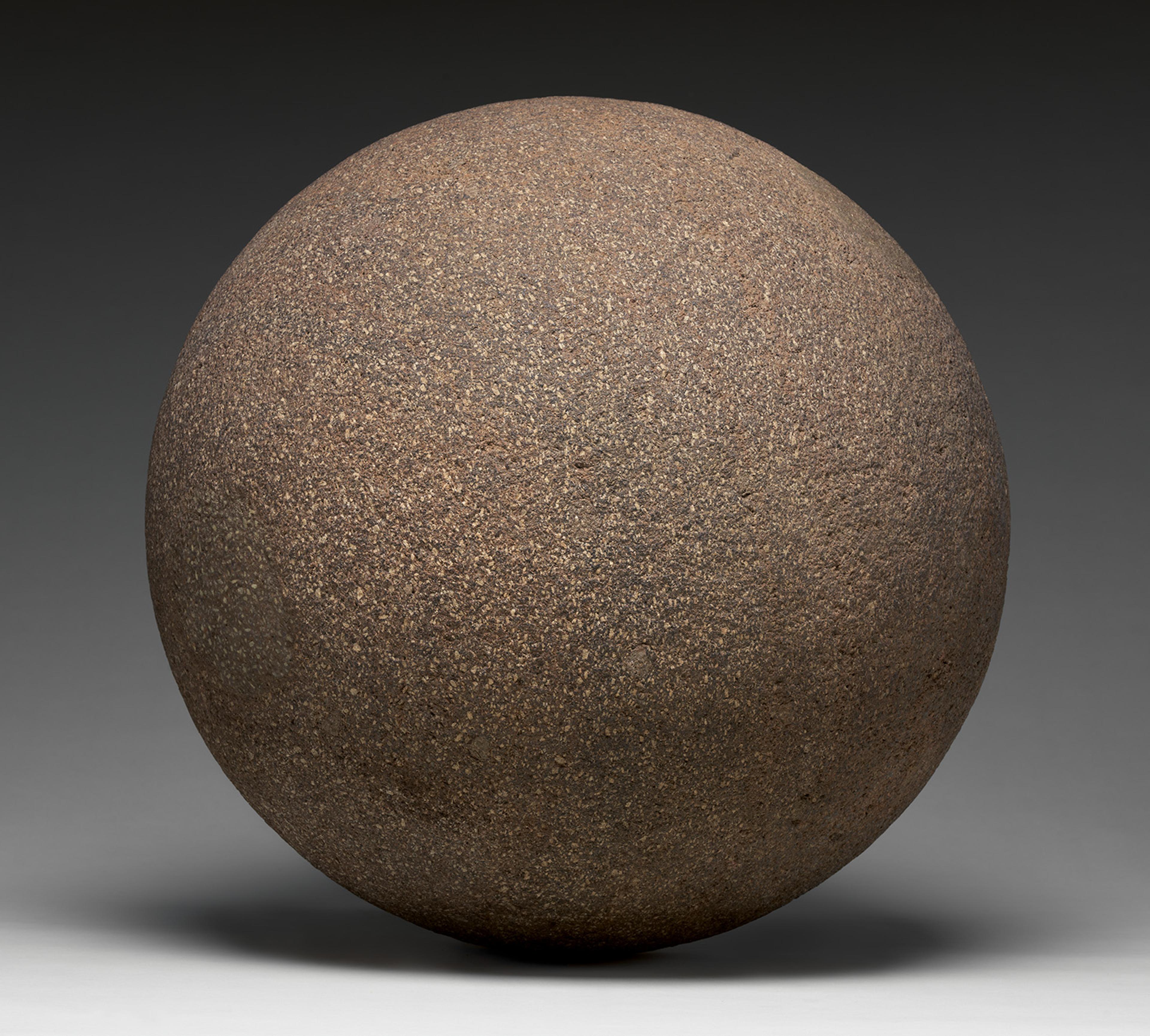 A large, weathered brown stone sphere rests on a gray surface, casting a soft shadow against a dark background